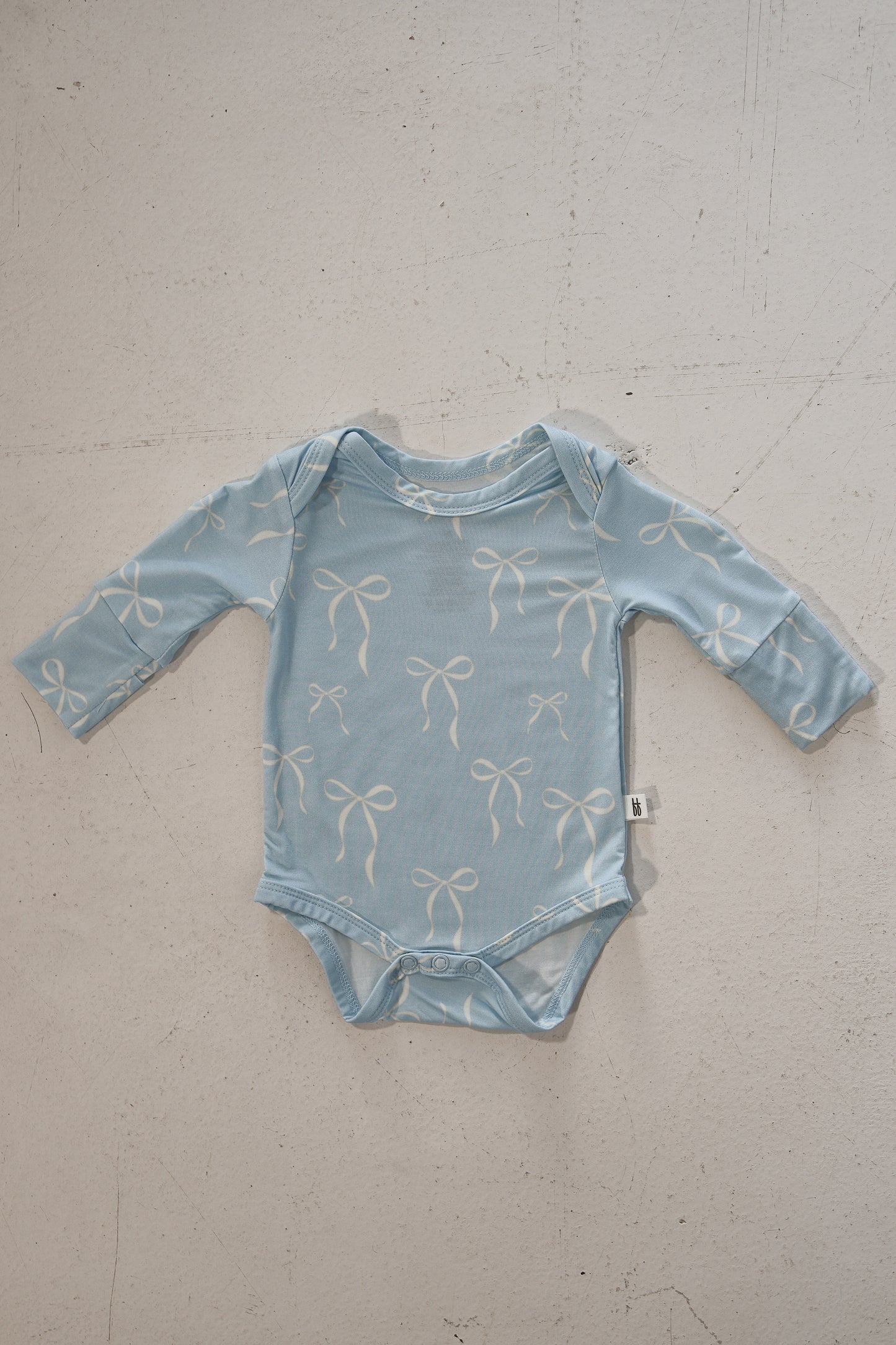 The forever french baby Long Sleeve Bamboo Snapsuit | Blue Bows is a hypoallergenic, light blue baby snapsuit with white bows, shown laid flat on a textured surface with a small white side tag.