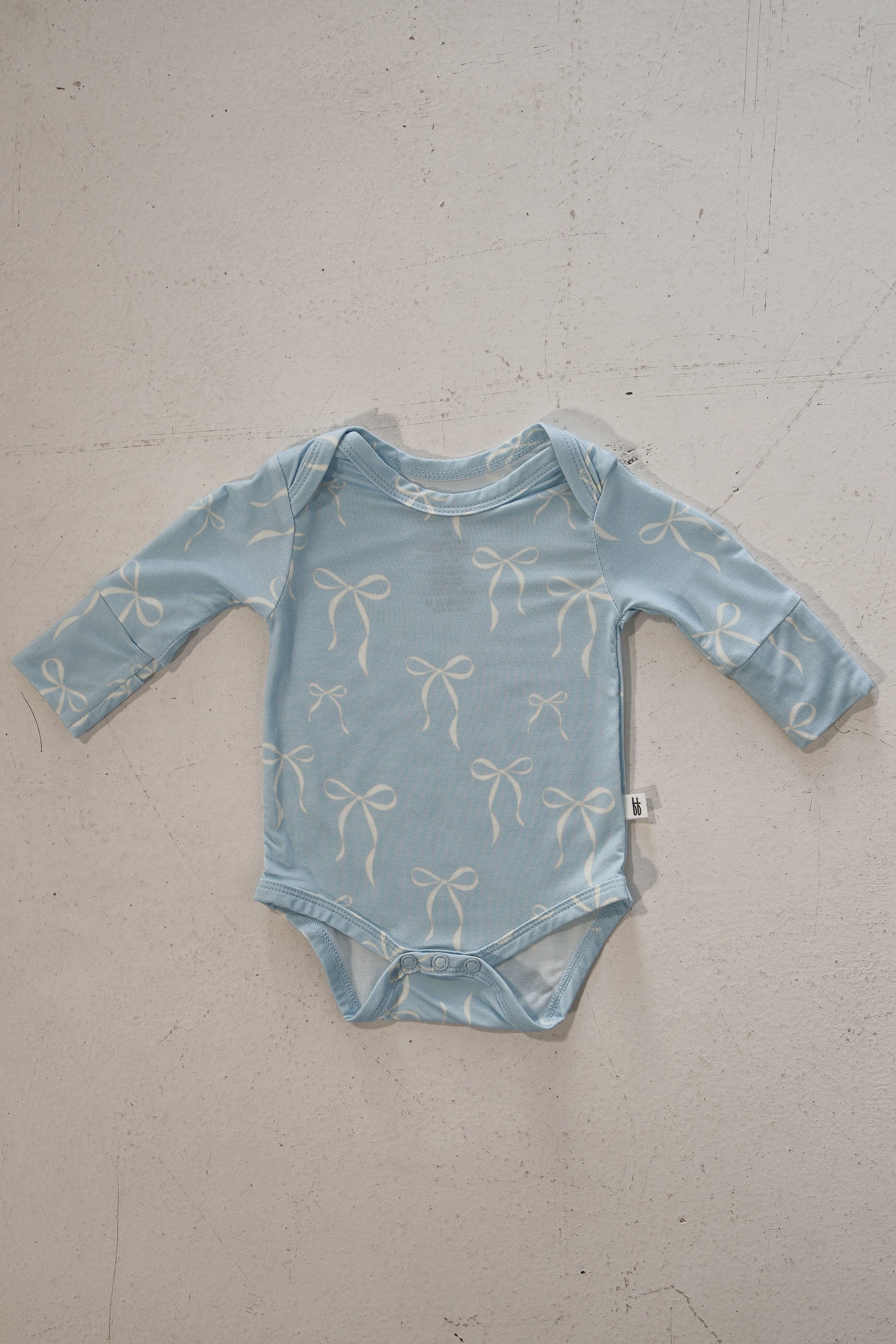 The forever french baby Long Sleeve Bamboo Snapsuit | Blue Bows is a hypoallergenic, light blue baby snapsuit with white bows, shown laid flat on a textured surface with a small white side tag.