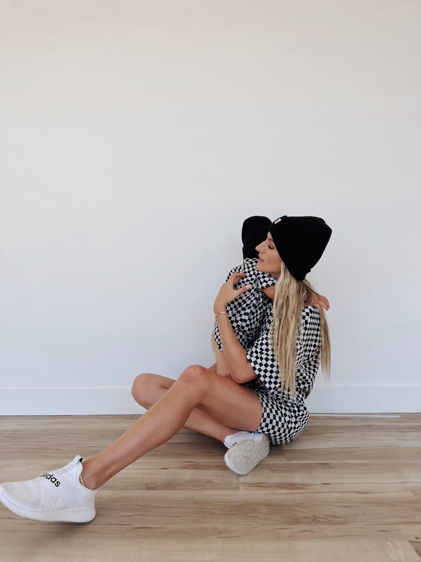 A woman sits on a wooden floor with her legs outstretched, wearing a black beanie, black and white checkered outfit, and white sneakers. She embraces a young child dressed in the Short Sleeve Ribbed Romper in Black & Grey Checkerboard from forever french baby, both sporting black beanies against a plain wall background.