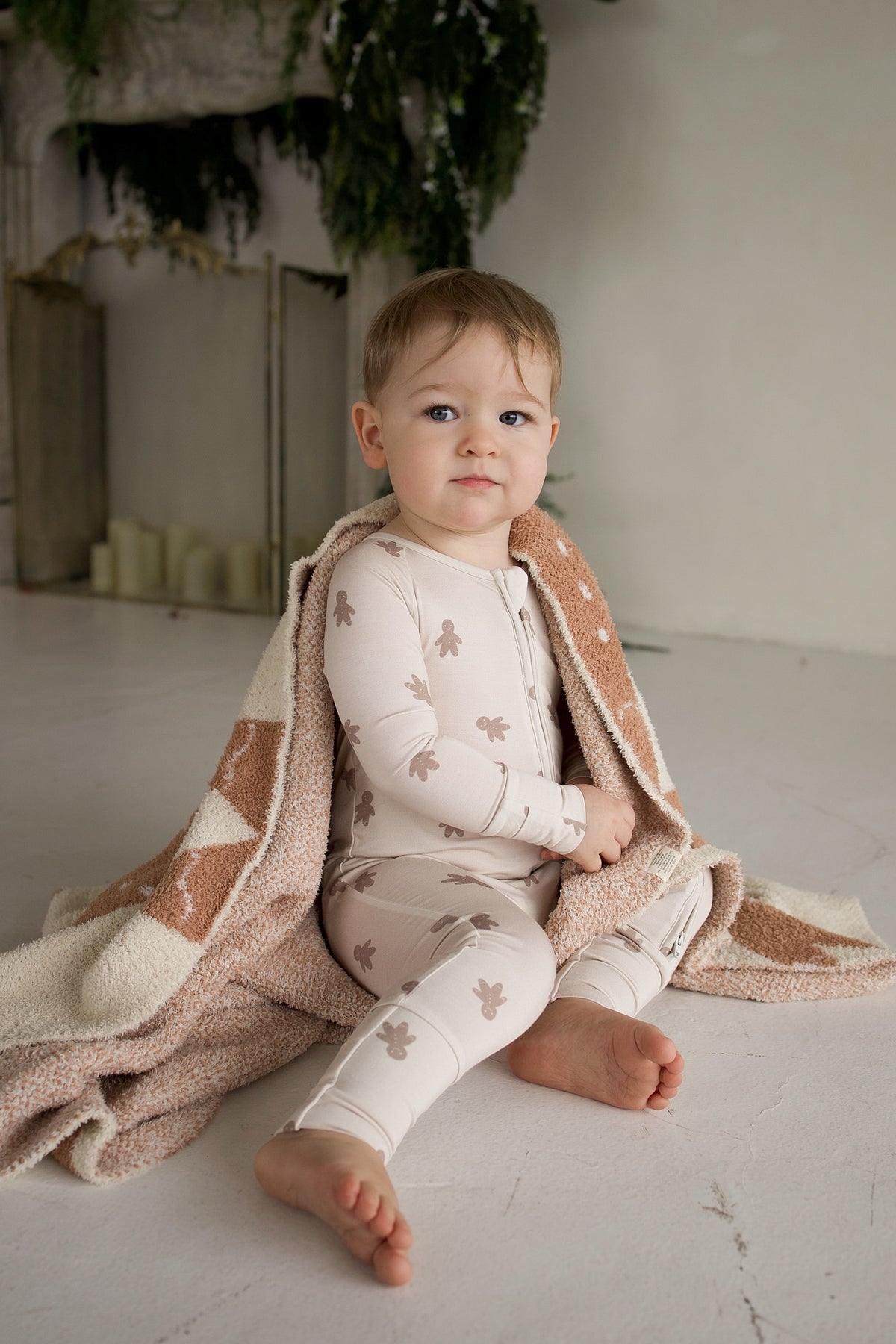 A baby in Bamboo Zip Pajamas | Gingersnap patterned with gingerbread men, wrapped in a patterned throw blanket.