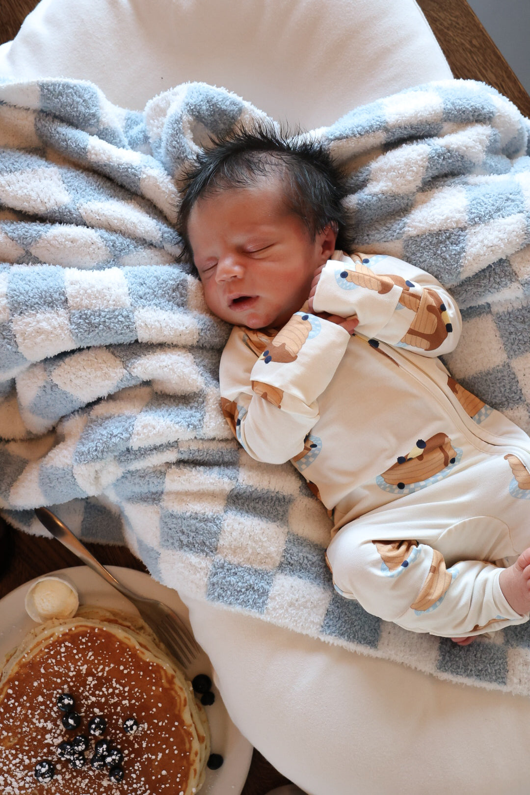 A newborn sleeps in forever french baby's Bamboo Zip Pajamas | Sunday Stack, featuring a bear-print onesie, on a blue and white checkered blanket with pancakes and blueberries nearby.