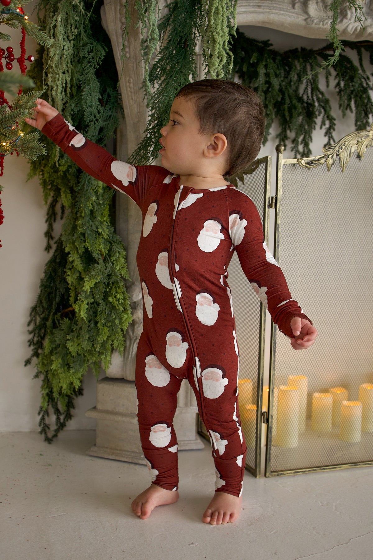 A toddler wearing Bamboo Zip Pajamas | Vintage Santa reaches toward a decorated Christmas tree.
