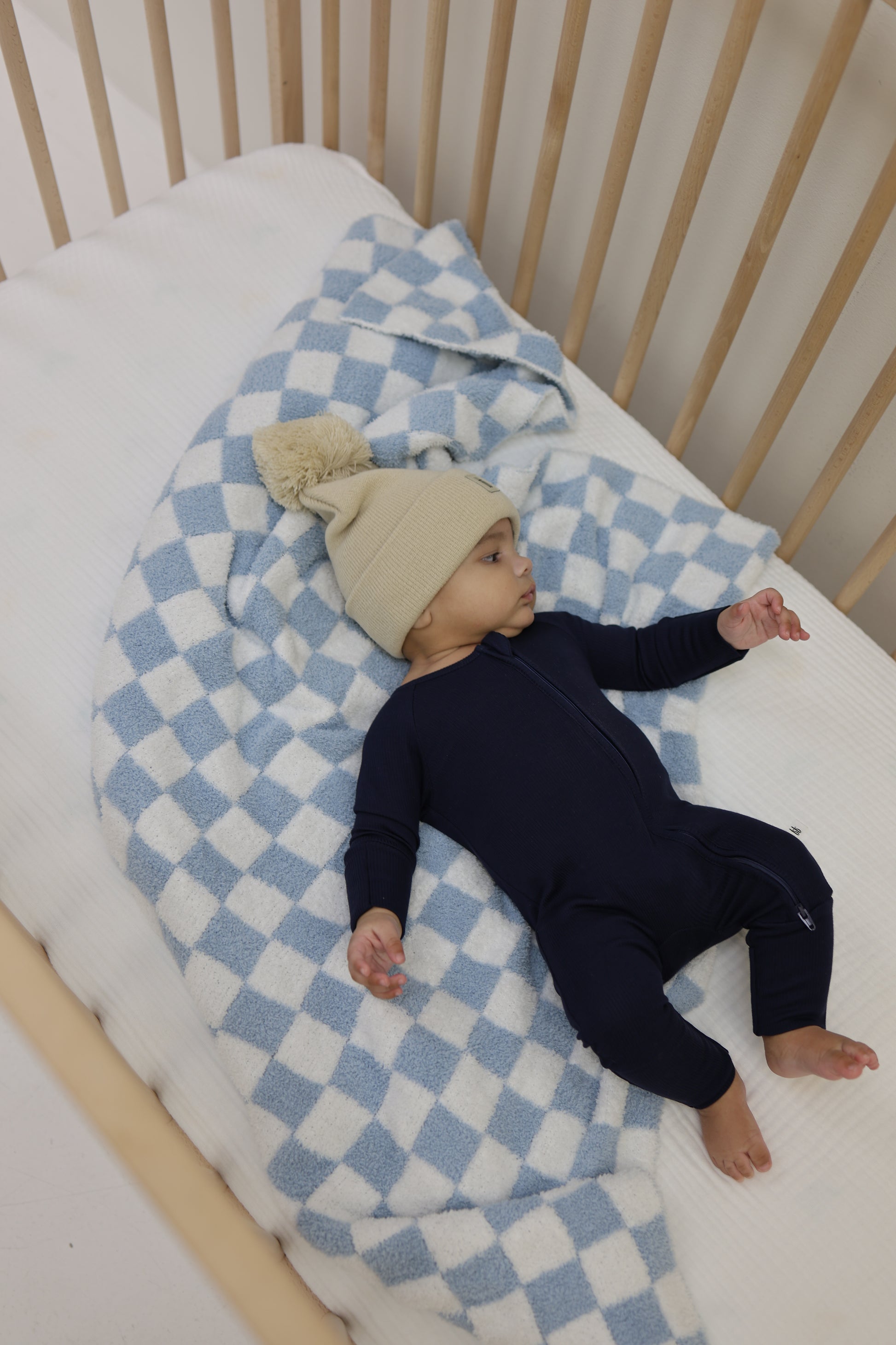 A baby in forever french baby's Bamboo Zip Pajamas | Blueberry Ribbed lies on a blue and white checkered blanket in a wooden crib, wearing a beige knit hat and looking off to the side.