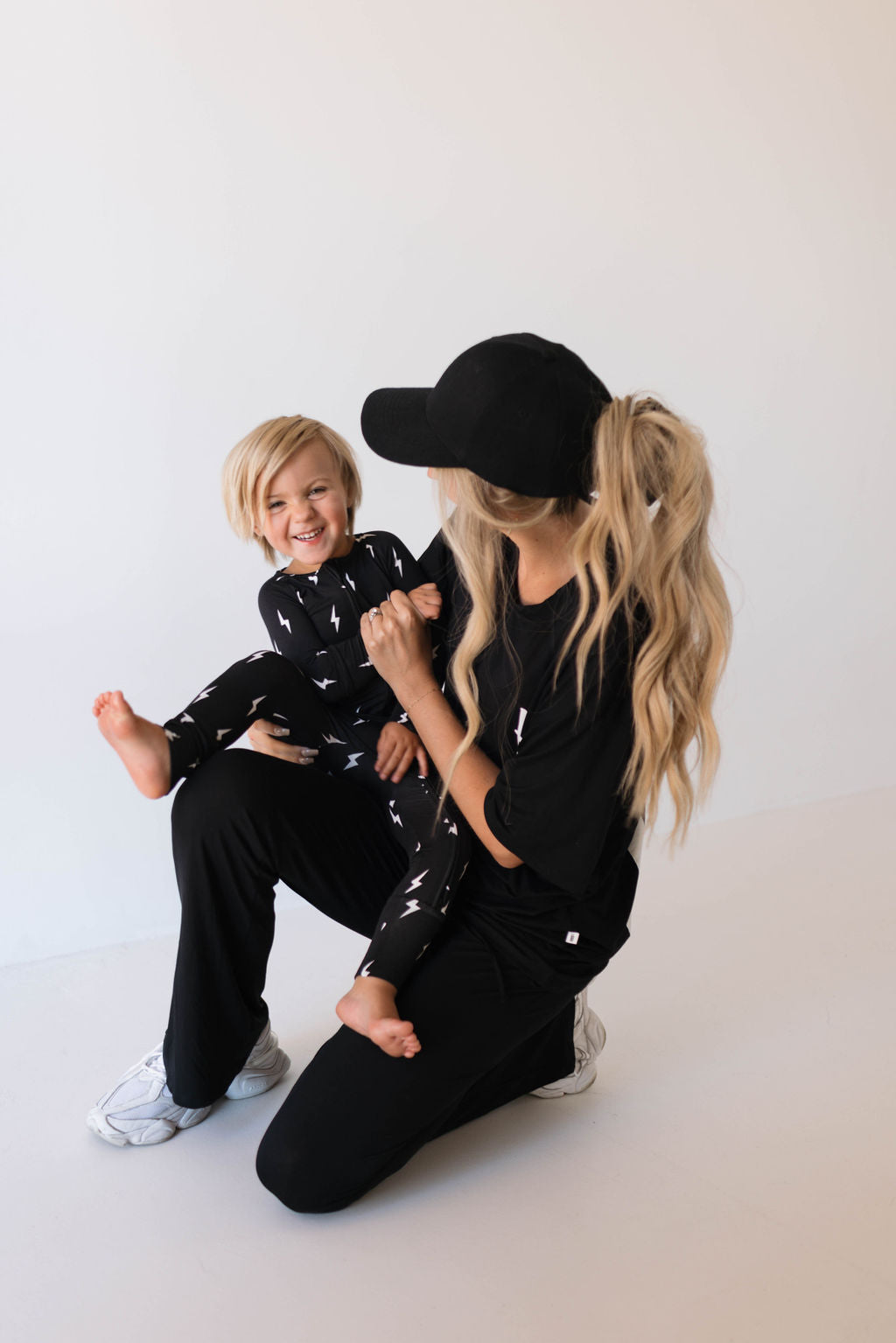 A woman with long blonde hair in a ponytail, wearing the forever french baby Short Sleeve Women's Bamboo Lounge Set | Black ff Signature, kneels on the floor holding a laughing child with blond hair. The child is dressed in matching black hypo-allergenic bamboo clothing adorned with white lightning bolt patterns. They are both smiling.