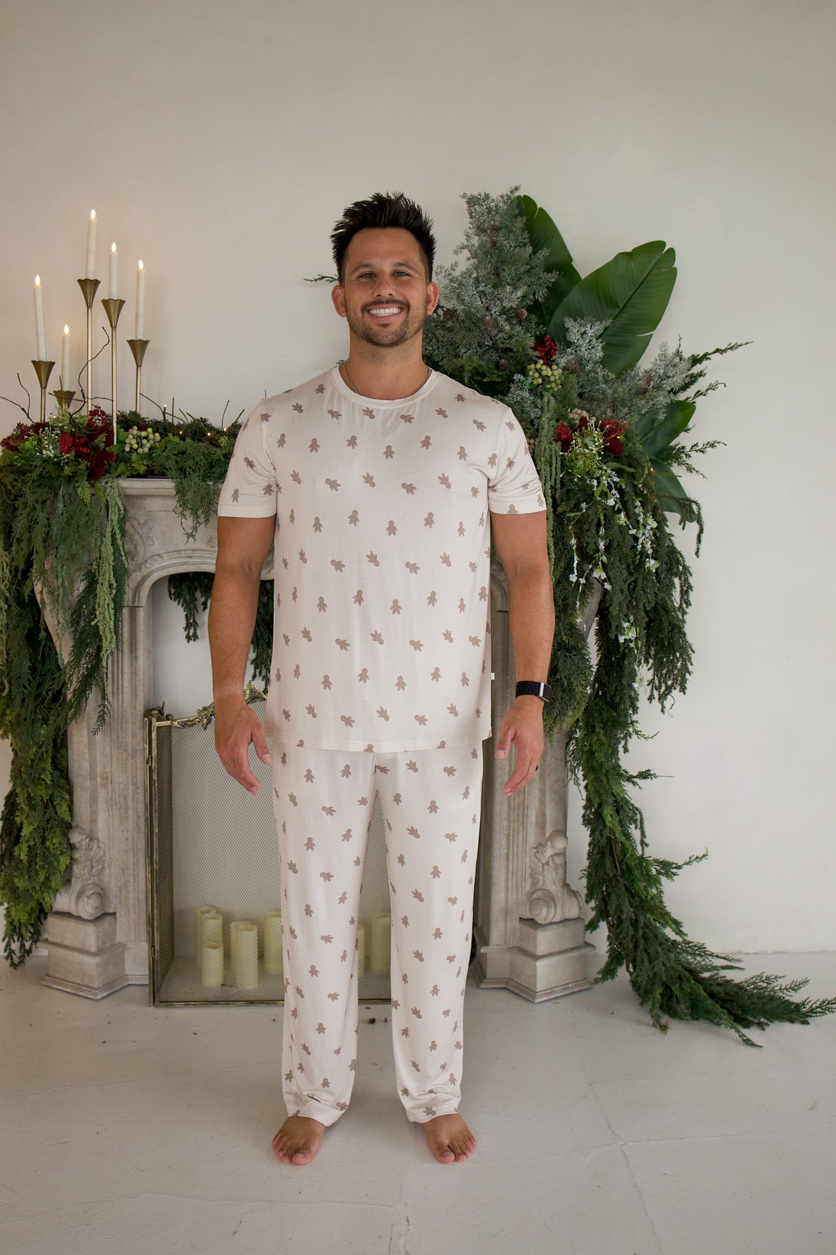 A man wearing a forever french baby Men’s Short Sleeve Pajamas | Gingersnap set, featuring a short-sleeved top and pants with a brown gingerbread man print.