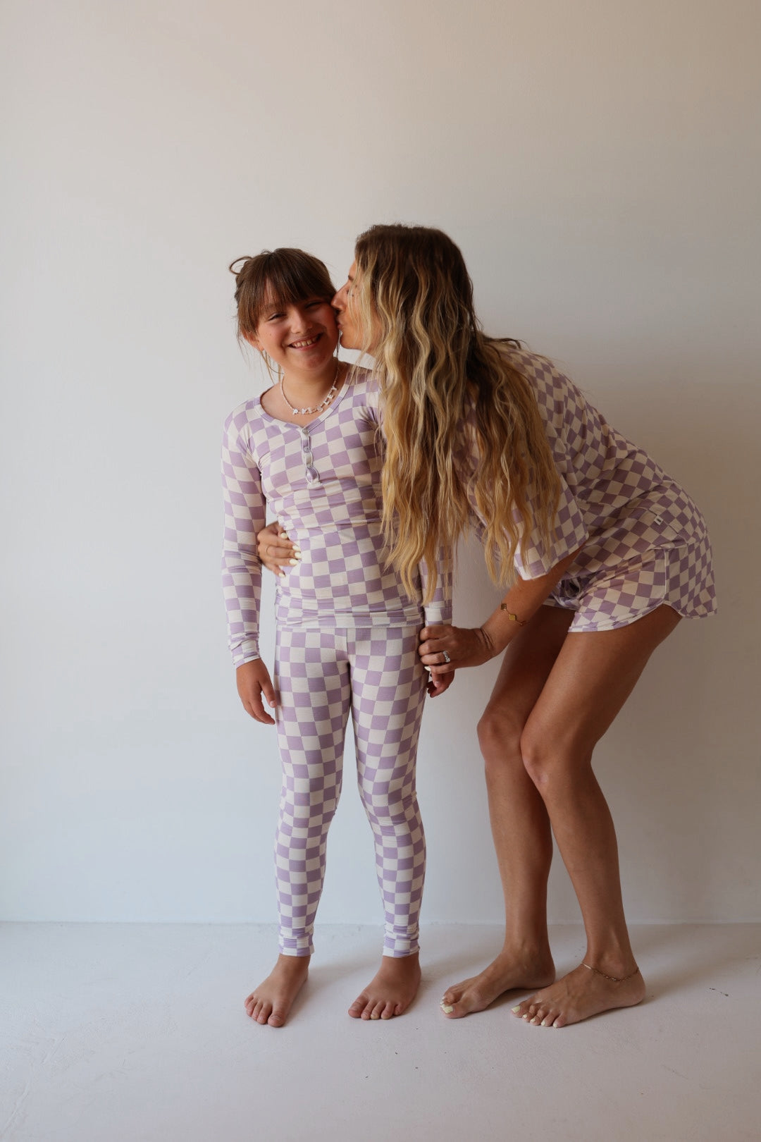 A woman kisses a smiling young girl on the cheek. Both are barefoot, wearing matching forever french baby Bamboo Two Piece Pajamas in Grape Soda, standing against a plain white background.