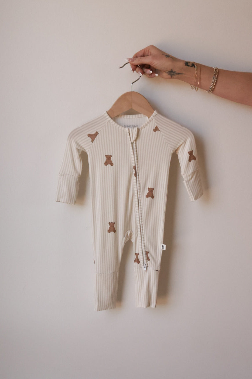 A hand holds up the forever french baby Bamboo Zip Pajamas | Teddy—a beige, long-sleeved, footed one piece with brown teddy bear prints and stripes, shown against a light wall—soft and hypoallergenic for babies.