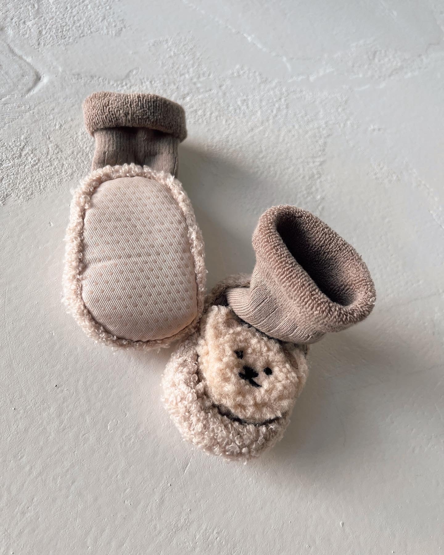 A pair of beige baby mittens crafted from soft, plush material with ribbed wrist cuffs. The mittens feature an adorable bear face design on the front, adding a cute touch reminiscent of the Teddy Bear Slipper Socks by forever french baby. They are displayed on a light-colored surface.