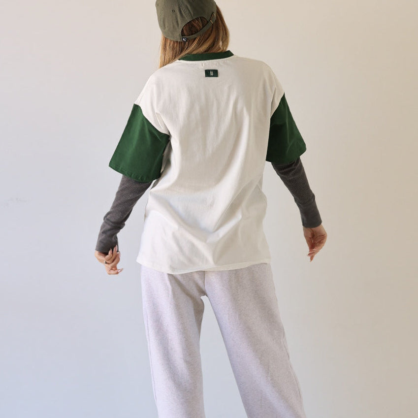 Person stands with their back to the camera, wearing a forever french baby Adult Cotton Tee | Lucky (white and green) over a gray long-sleeve shirt, white sweatpants, sneakers, and a gray cap. Background: plain off-white wall.