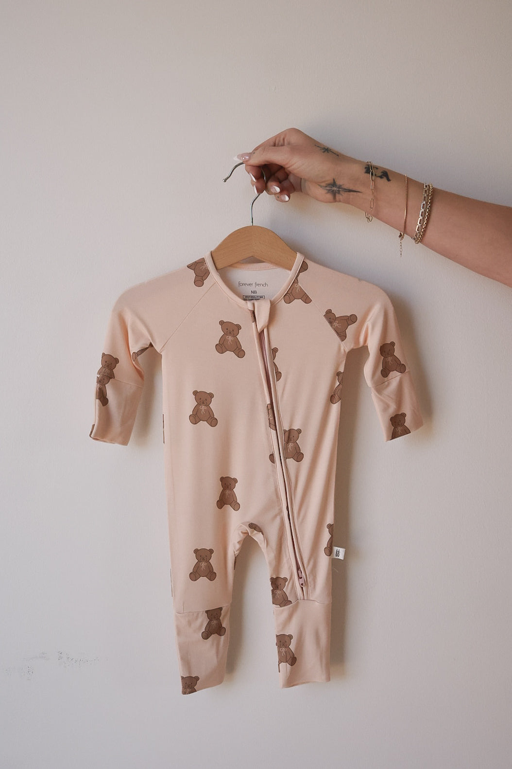 A hand holds forever french baby's Bamboo Zip Pajamas | Lottie—a beige bamboo romper with brown teddy bear print, perfect as breathable baby sleepwear—against a plain light background.
