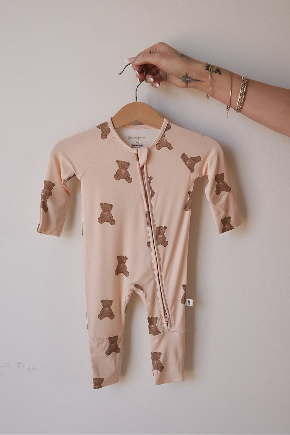 A hand holds forever french baby's Bamboo Zip Pajamas | Lottie—a beige bamboo romper with brown teddy bear print, perfect as breathable baby sleepwear—against a plain light background.