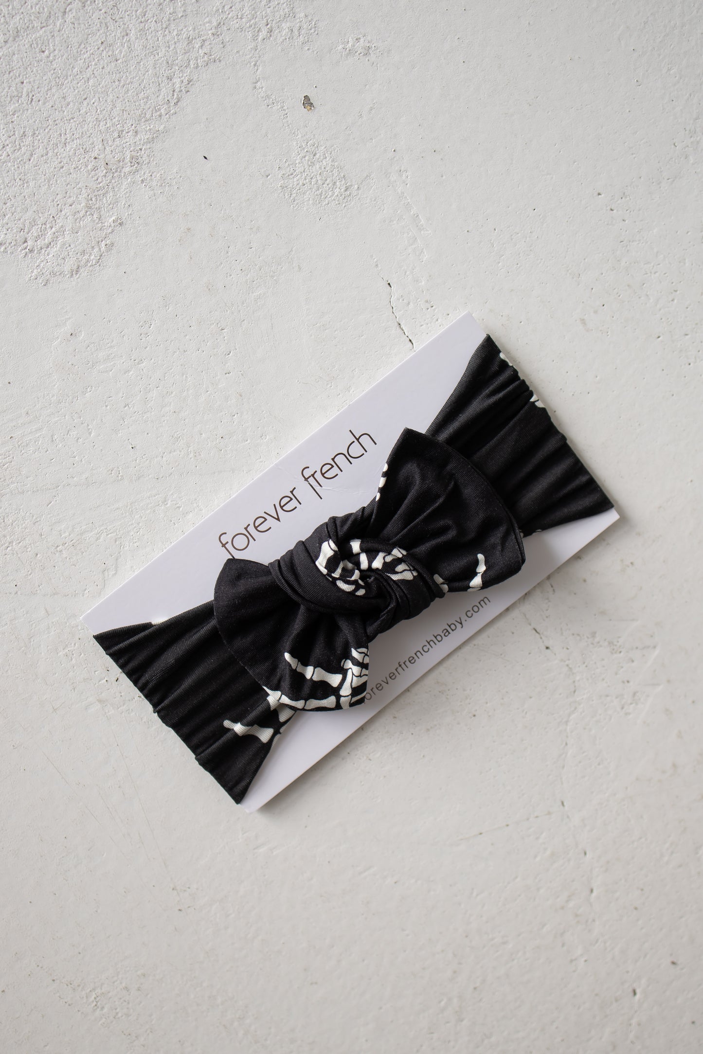 A black pleated fabric baby bow headband featuring white abstract accents, named the Bamboo Head Wrap | Midnight Skelly by forever french baby, displayed on a “forever french” branded card on a white textured surface.