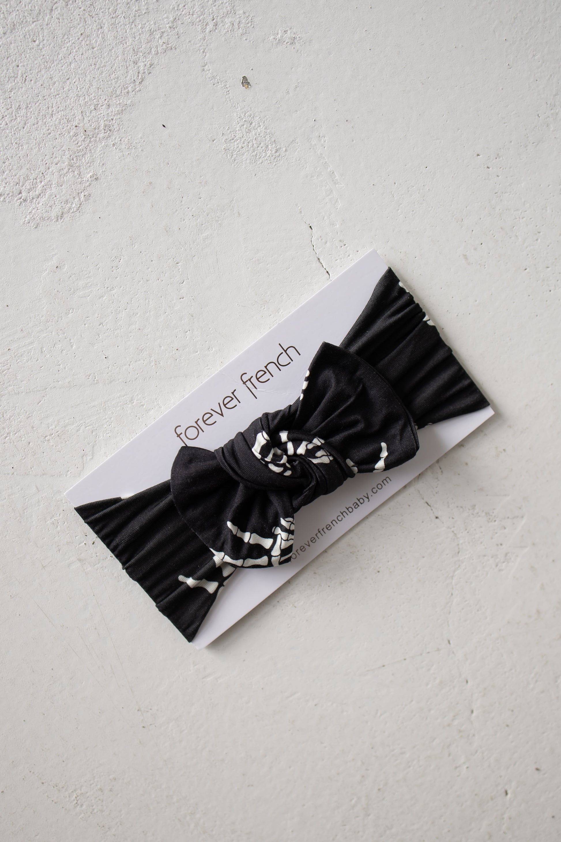 A black pleated fabric baby bow headband featuring white abstract accents, named the Bamboo Head Wrap | Midnight Skelly by forever french baby, displayed on a “forever french” branded card on a white textured surface.