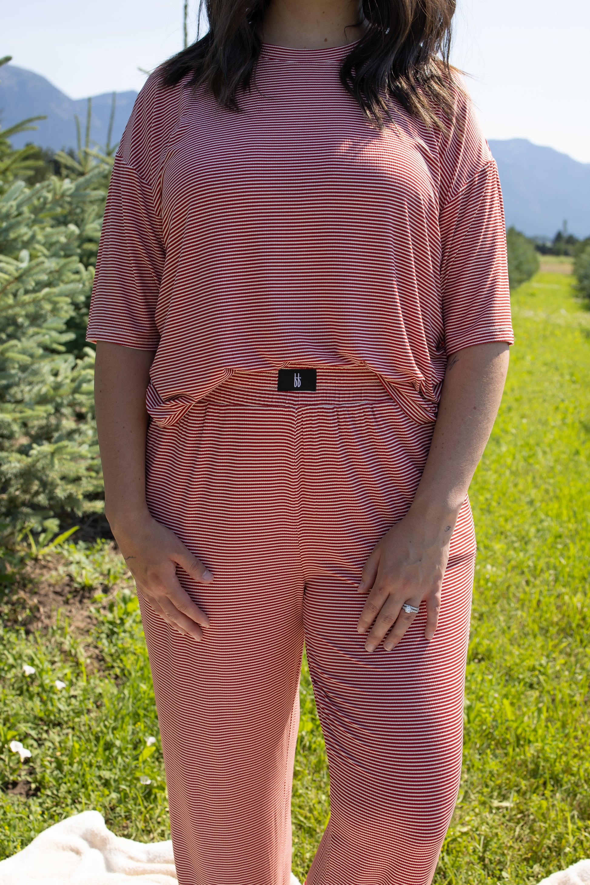 A woman stands outdoors on grass wearing the forever french baby Women's Boxy Tee Lounge Set in Peppermint Stripe Ribbed. The matching short-sleeve top and pants are crafted from breathable loungewear, with her hands resting by her sides as mountains and trees appear in the background.