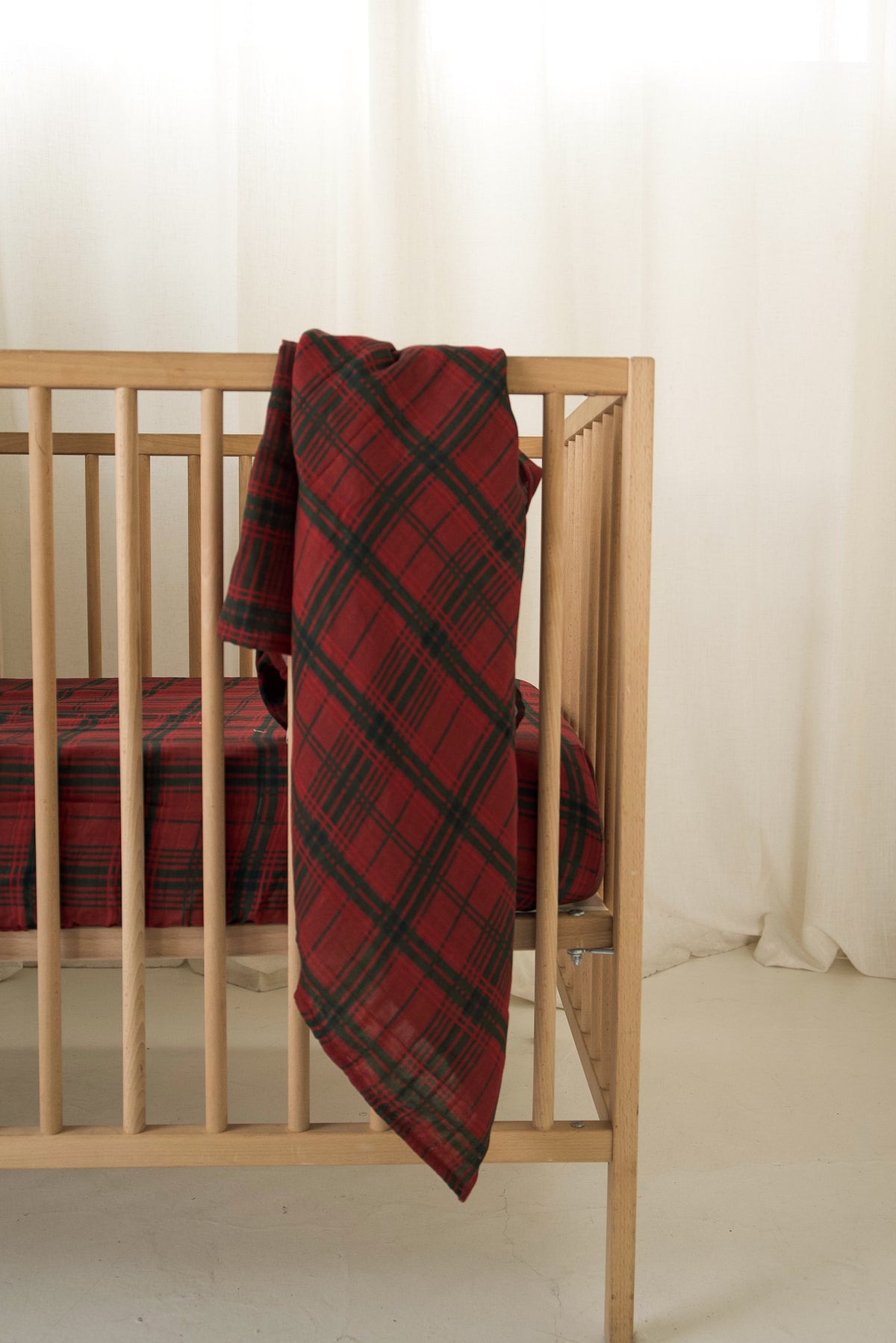 Forever French Baby Muslin Swaddle | Christmas Plaid blanket draped over the side of a wooden crib with a matching plaid sheet inside.