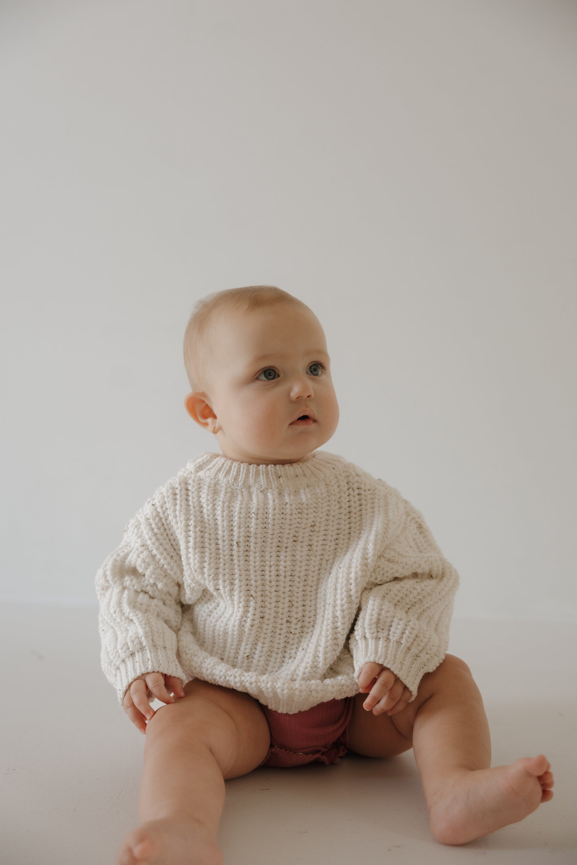 A light-skinned, short-haired baby sits on the floor against a plain, light background wearing the forever french baby Child Knit Sweater | Cloud Confetti and pink shorts.