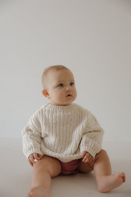 A light-skinned, short-haired baby sits on the floor against a plain, light background wearing the forever french baby Child Knit Sweater | Cloud Confetti and pink shorts.