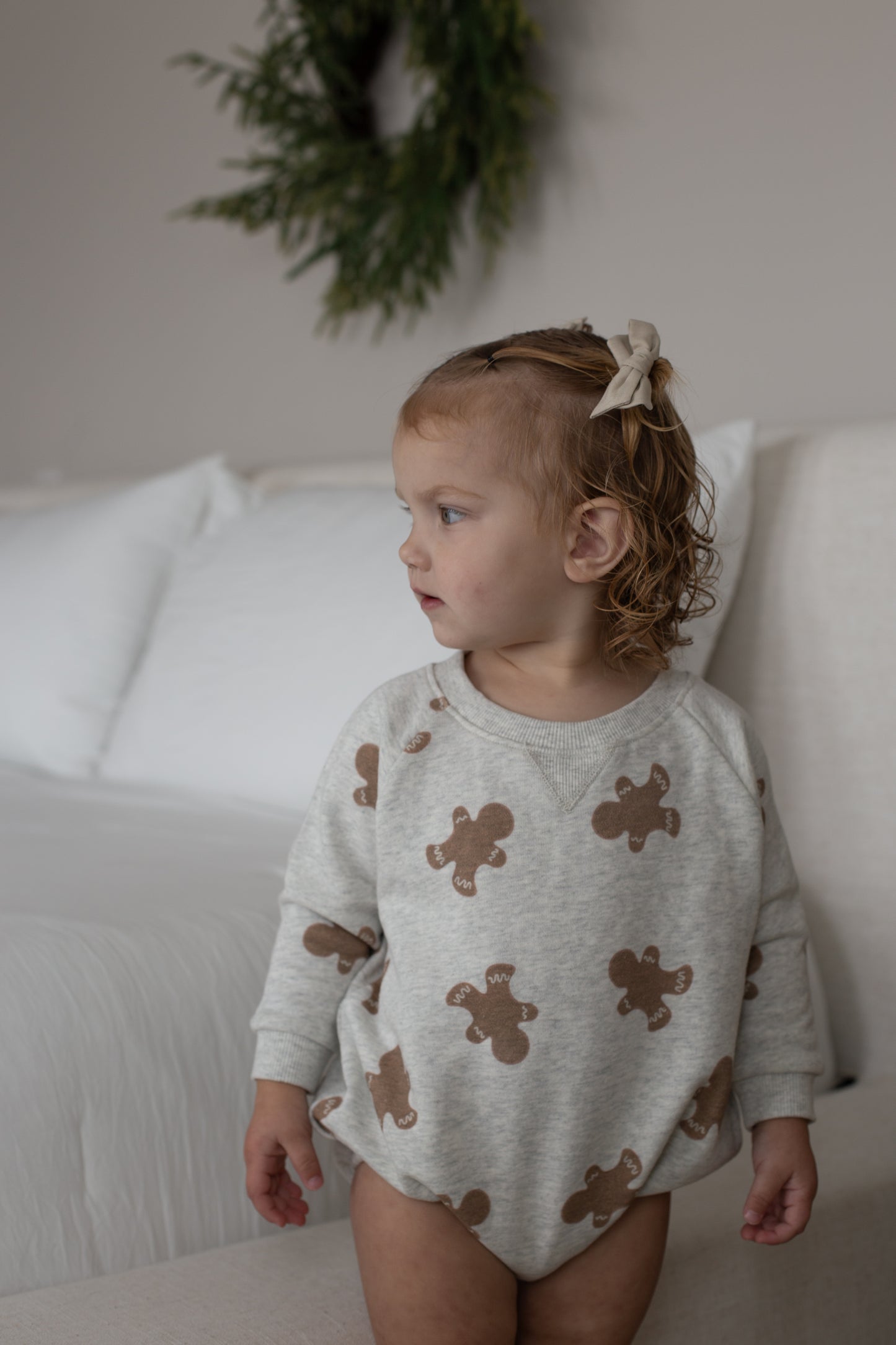 A young child with curly hair and a beige bow stands by a bed, wearing the forever french baby Sweatshirt Romper | Gingerbread 2.0. A green wreath decorates the wall in the background.