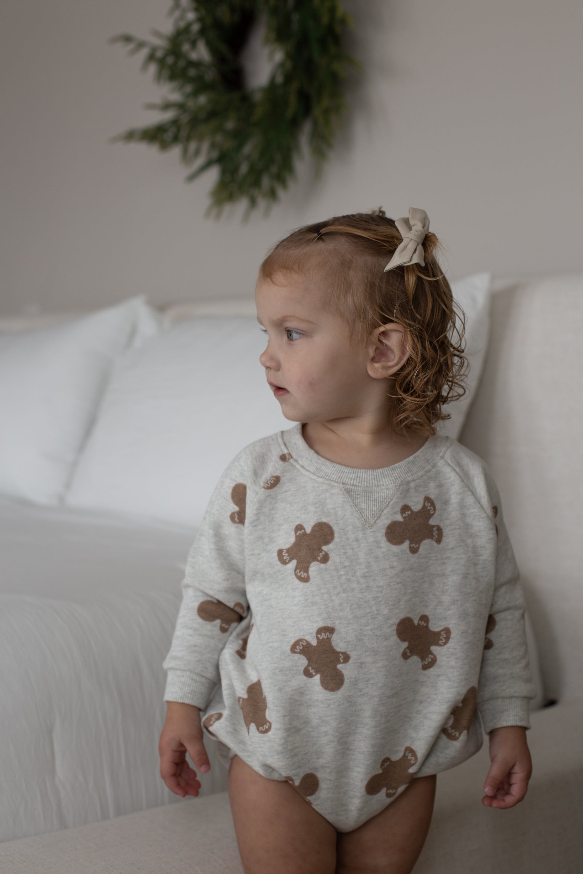 A young child with curly hair and a beige bow stands by a bed, wearing the forever french baby Sweatshirt Romper | Gingerbread 2.0. A green wreath decorates the wall in the background.
