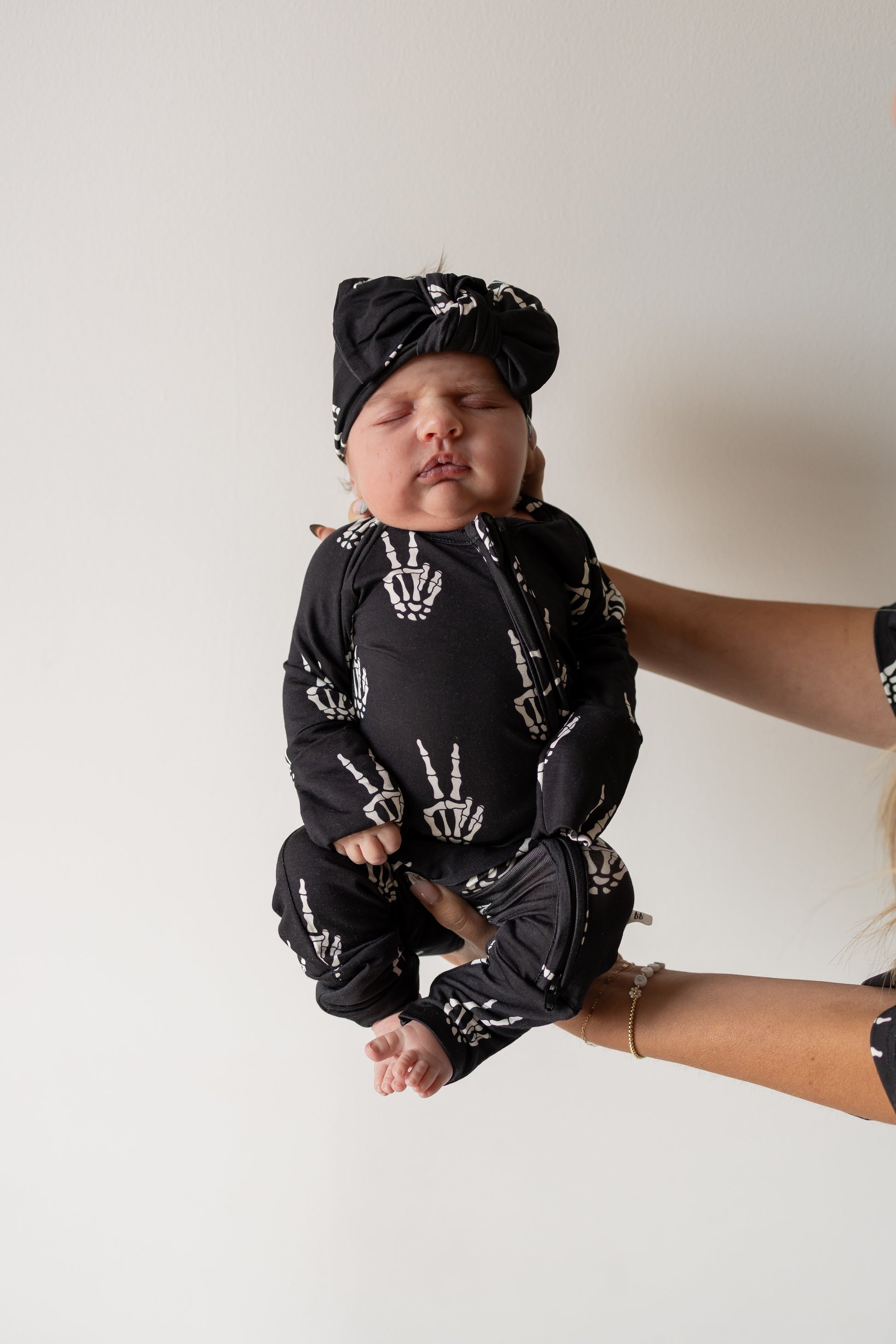 An adult holds up a baby dressed in forever french baby’s Bamboo Zip Pajamas | Midnight Skelly—black, skeleton-themed pajamas made from soft, hypo-allergenic fabric—against a plain white background.