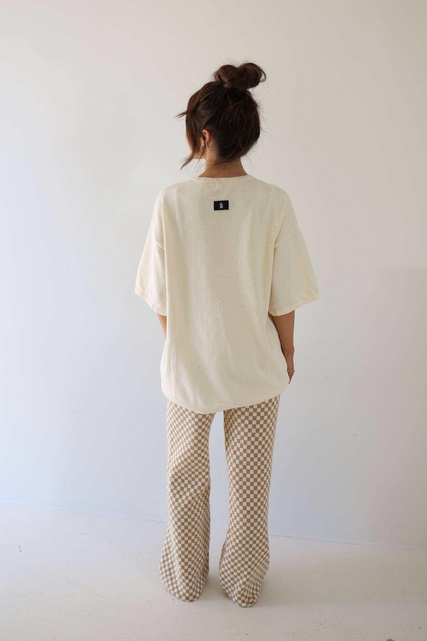 A woman with long brown hair in a messy bun stands against a plain white wall, wearing the forever french baby Women's Knit Pant Set | Croissants—an oversized cream top and beige checkered pants—in a minimal setting.