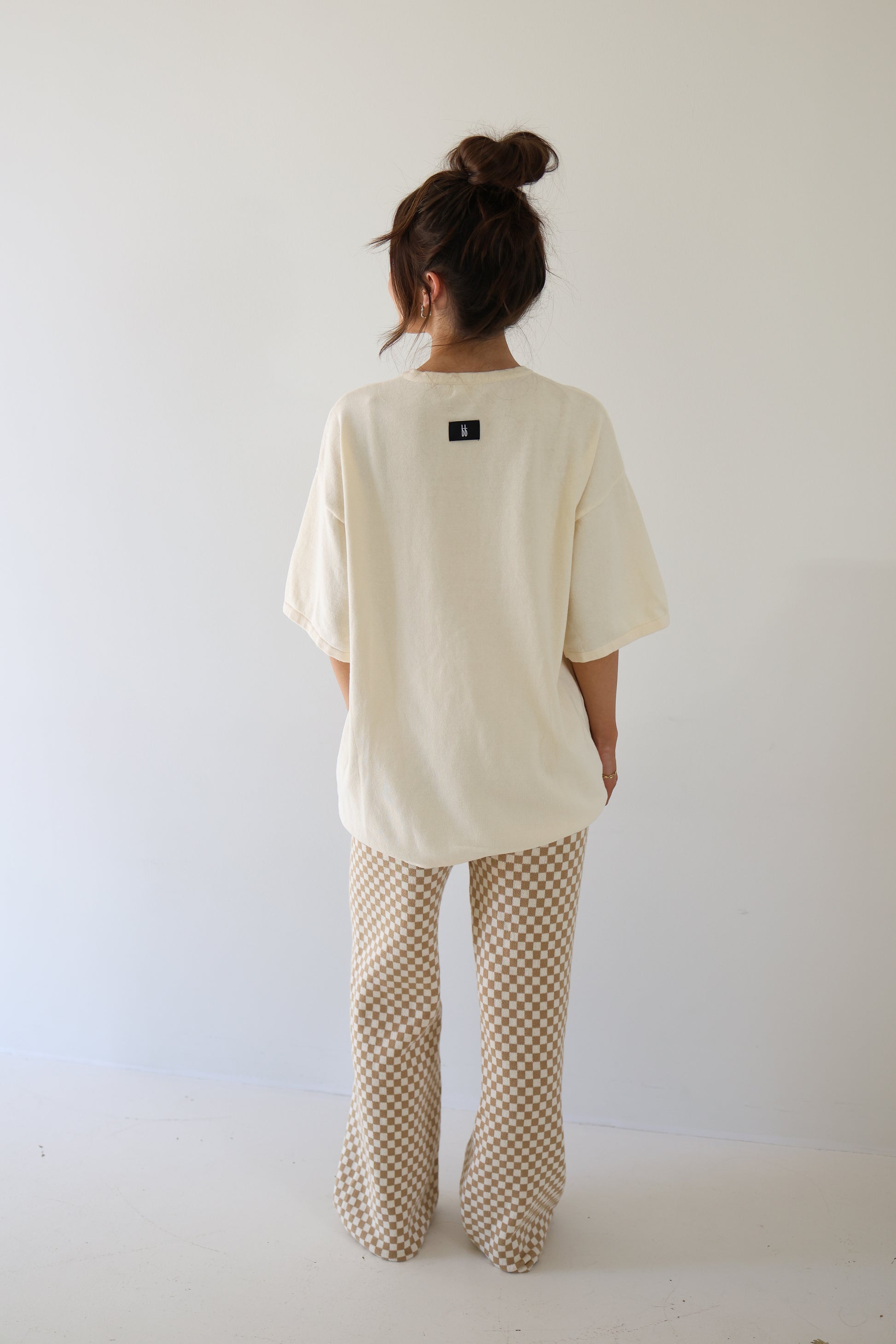 A woman with long brown hair in a messy bun stands against a plain white wall, wearing the forever french baby Women's Knit Pant Set | Croissants—an oversized cream top and beige checkered pants—in a minimal setting.