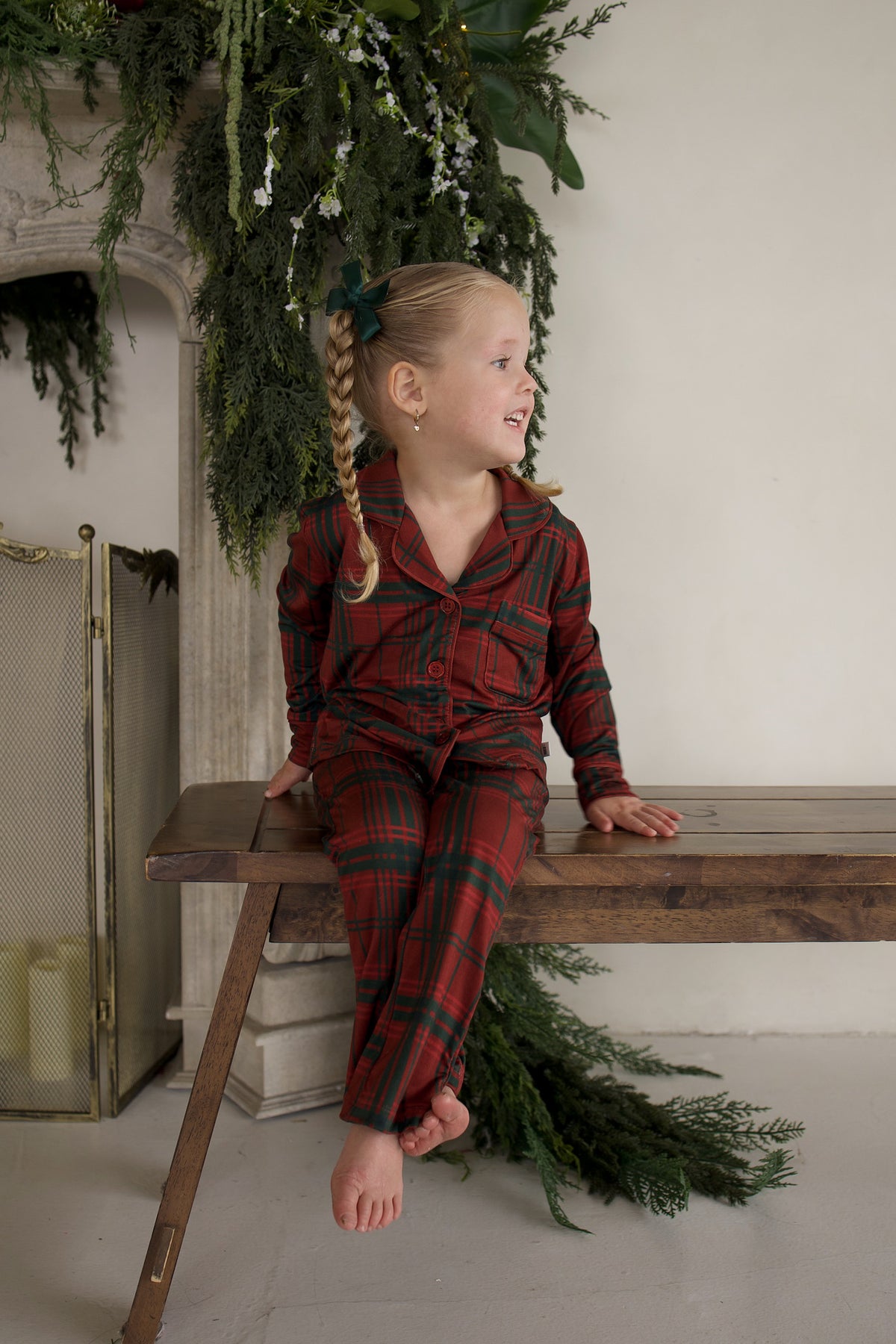 A young girl sits on a wooden bench wearing a Girl’s Bamboo Button Up Lounge Set | Christmas Plaid in a red and green long-sleeved plaid pattern.