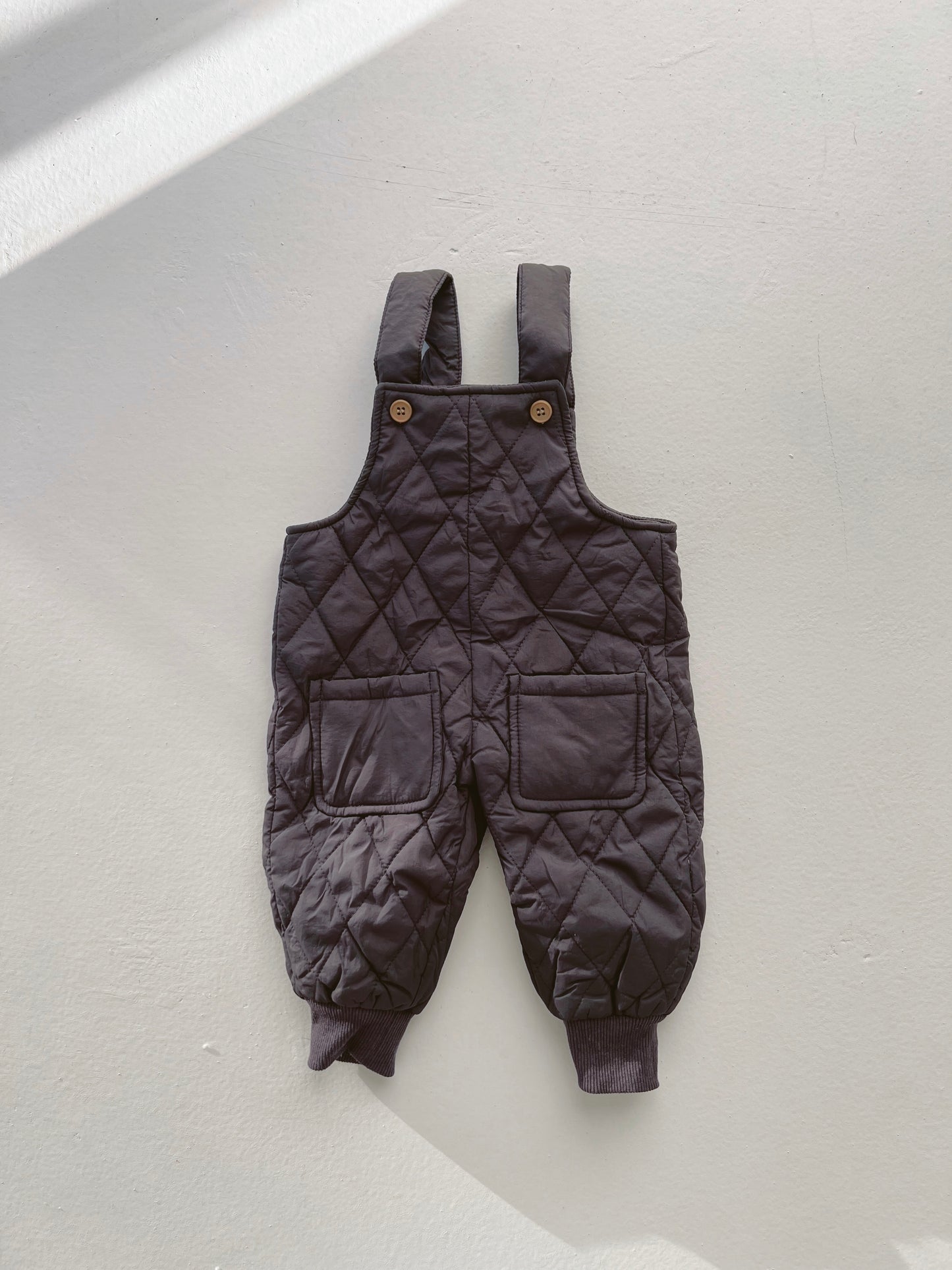 The Charcoal Snow Bibs by forever french baby are a pair of extra-warm, black quilted overalls featuring thick straps with wooden buttons at the top, two front pockets, and ribbed ankle cuffs. They are displayed against a light background.