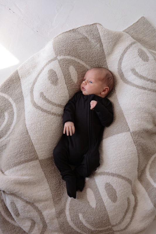 A newborn baby dressed in forever french baby's Bamboo Zip Pajamas in Black Ribbed lies on a cozy, hypo-allergenic blanket adorned with large smiley faces. The baby gazes to the side with a calm expression, enjoying the soft and light-colored background.