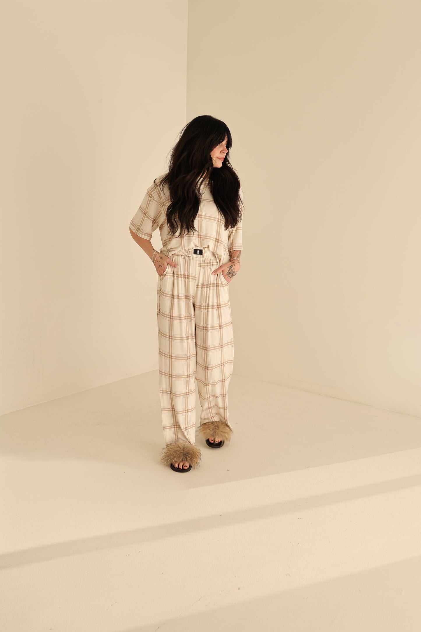A woman with long dark hair stands in a beige-toned room, wearing the forever french baby Women's Boxy Tee Lounge Set in Ellis Plaid, featuring wide-legged pants and furry slides, as she looks to the side with her hands in her pockets.
