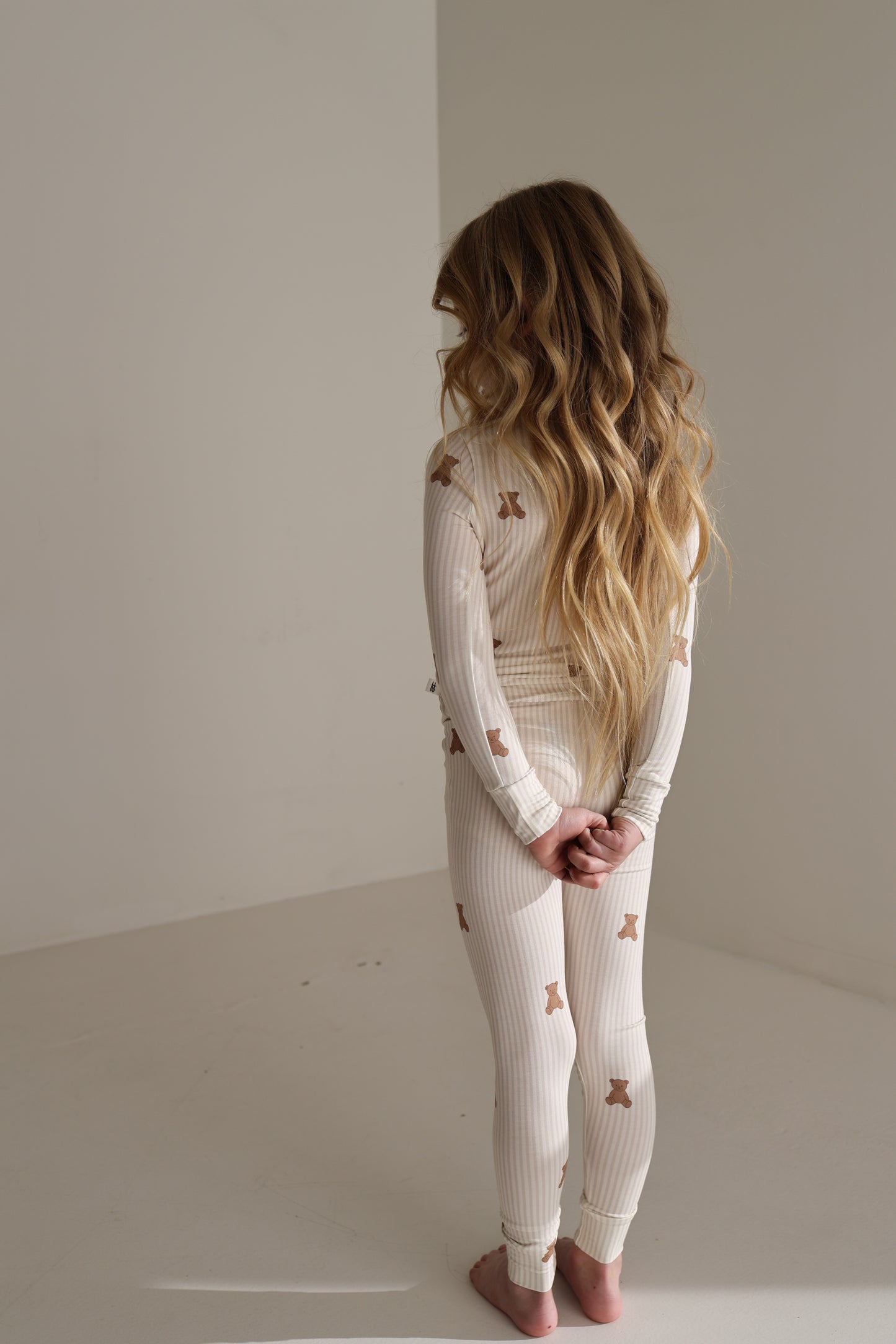 A young child stands barefoot in a white room, wearing forever french baby's Bamboo Two Piece Pajamas | Teddy—cream-colored with brown bear prints—hands clasped behind their back, long wavy hair flowing.