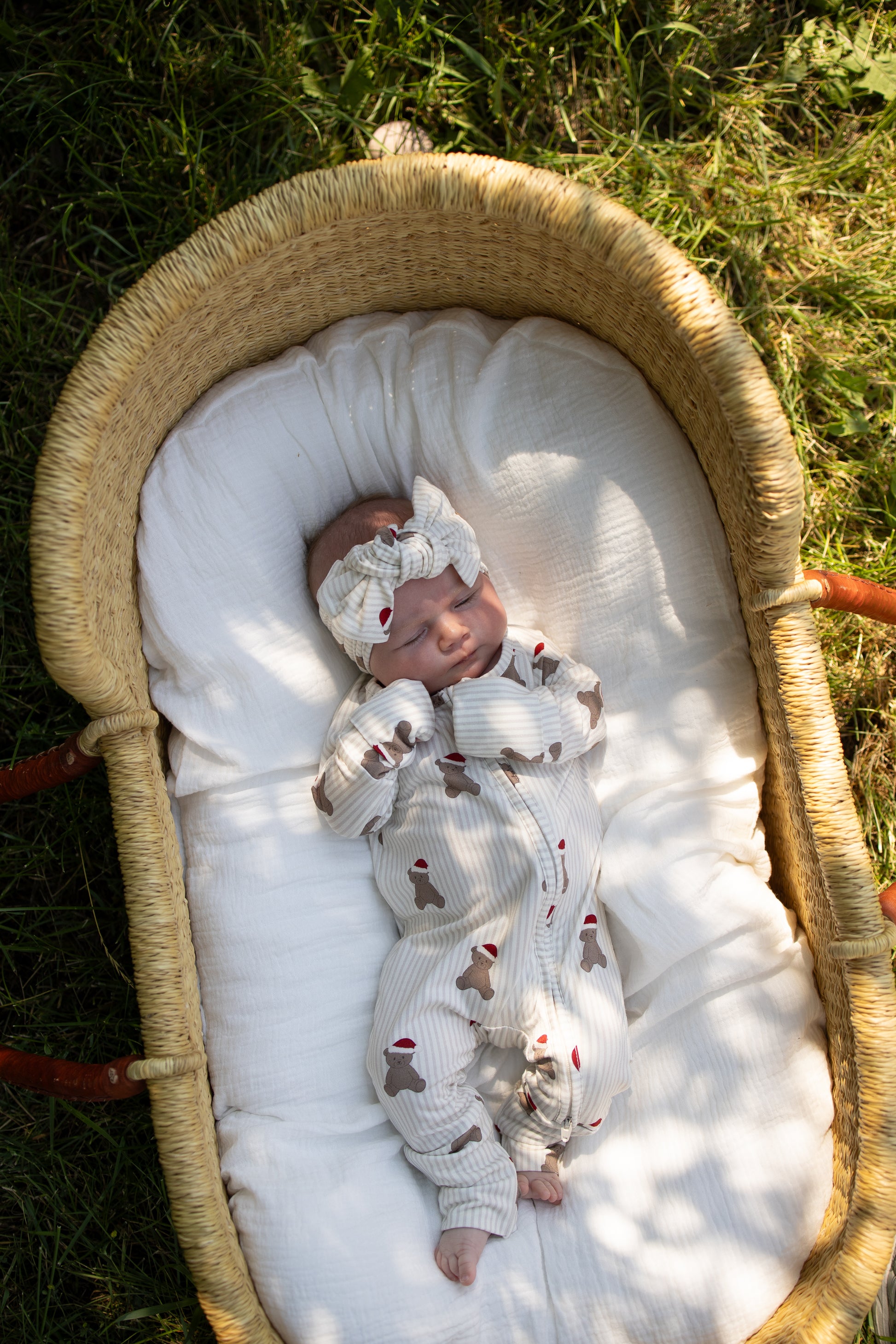 A newborn baby sleeps outdoors in a wicker bassinet, dressed in the forever french baby Bamboo Zip Pajamas | Santa Teddy—a matching cream outfit and headband adorned with brown dog patterns, made from hypoallergenic fabric—while lying on a soft white blanket in the sunlight.