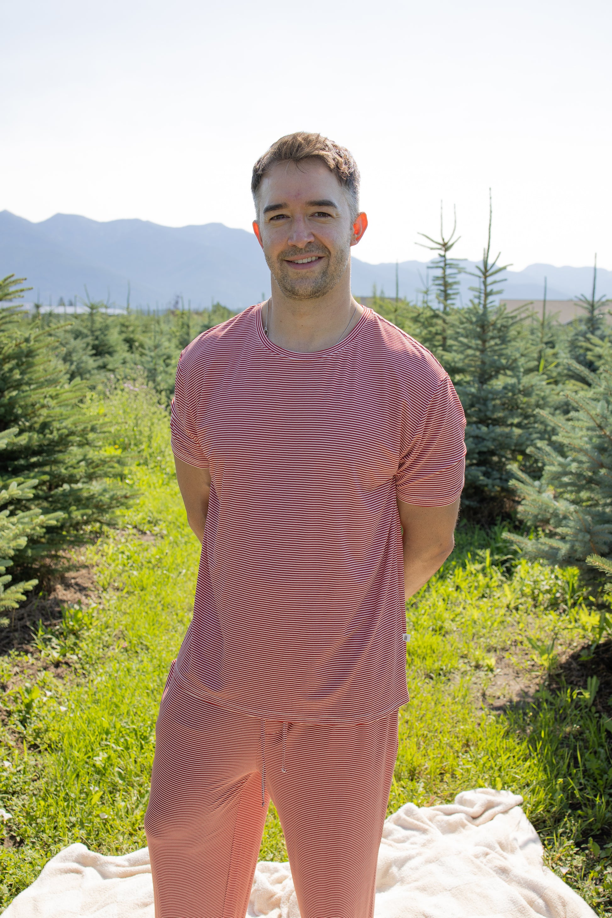 Among evergreen trees beneath a sunny mountain backdrop, a person stands outdoors on a light blanket wearing the Men's Short Sleeve Pajamas in Peppermint Stripe Ribbed by forever french baby. These hypo-allergenic pajamas combine stylish comfort with nature’s beauty.