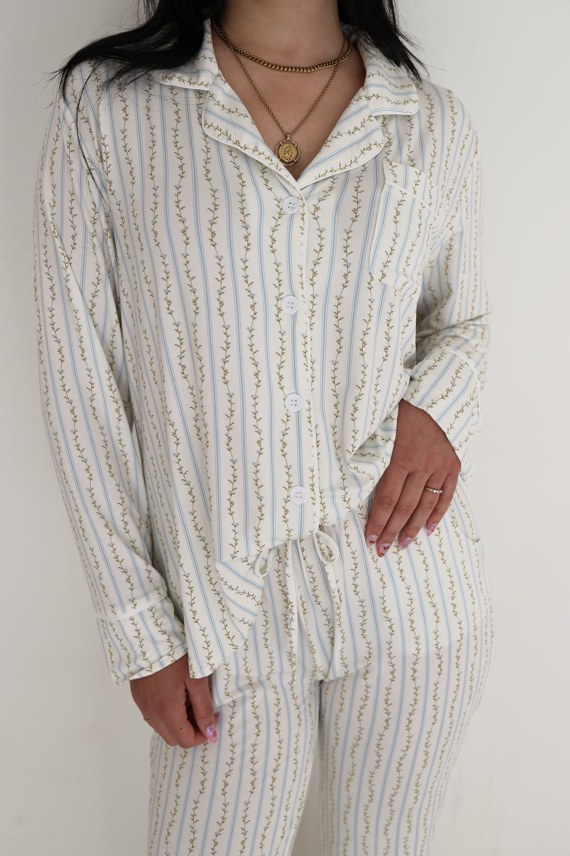 A woman wears the forever french baby Women's Bamboo Button Up Lounge Set in Dainty Vines, featuring a white base with gold vine stripes, accessorized with gold jewelry and posing with her hand on her hip. The set is hypo-allergenic bamboo.