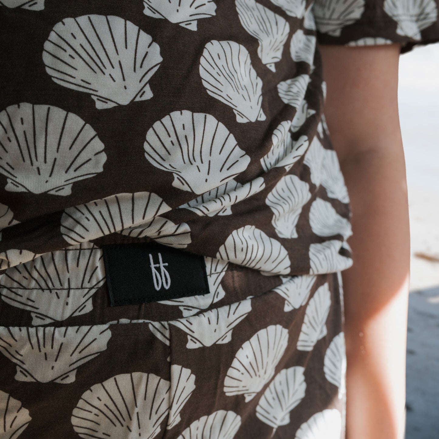Close-up of a child wearing the forever french baby Child Boxy Tee Lounge Set | Seashells, a breathable brown outfit with white seashell patterns and a black “bb” fabric label, standing near a sandy beach. The attire is hypo-allergenic.