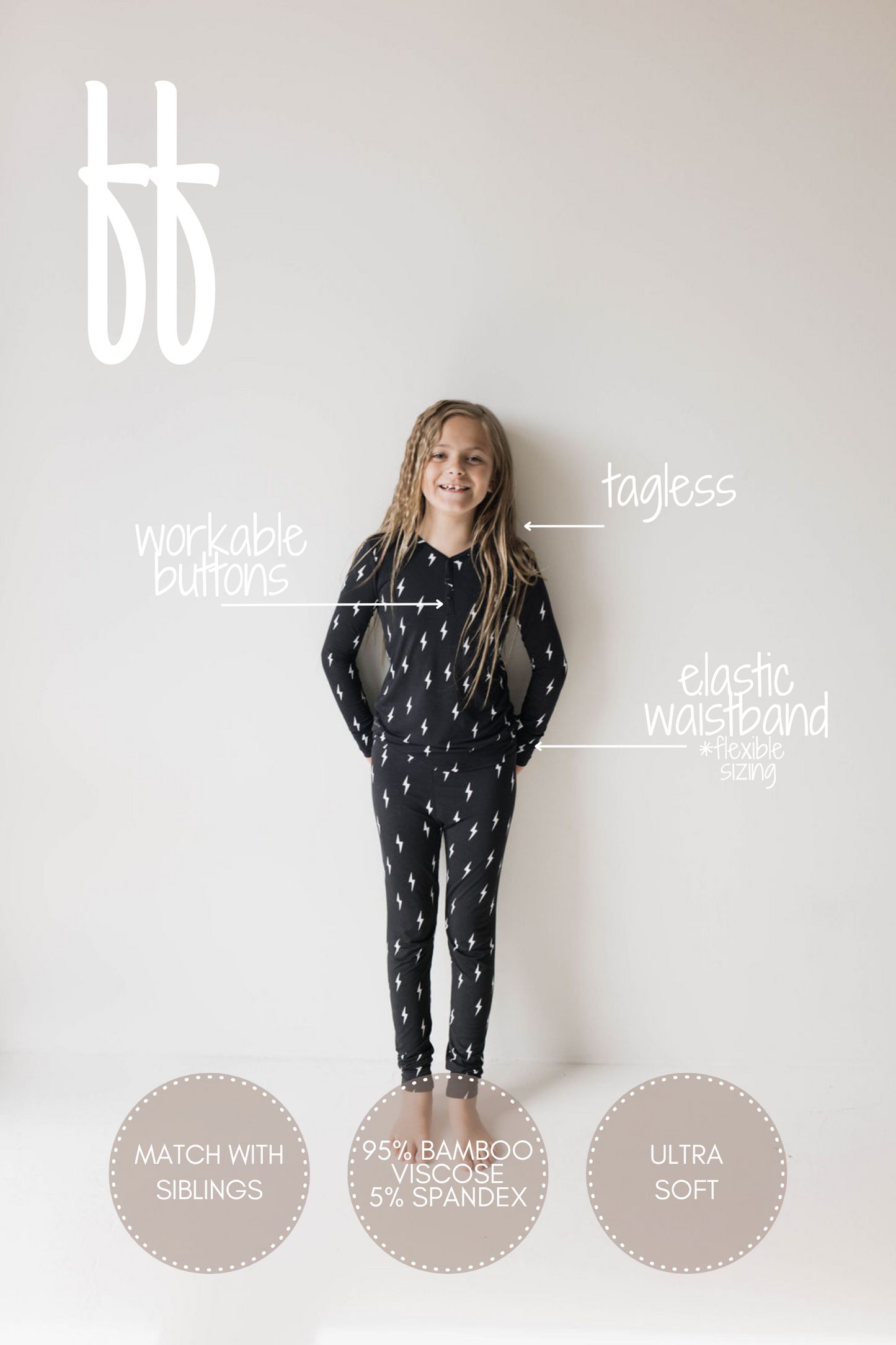 A young girl with long hair stands against a plain white background, wearing the "Bamboo Two Piece Pajamas | Midnight Lightning Bolt" by forever french baby. The ultra-soft black outfit showcases white lightning bolt patterns and features labels indicating "workable buttons," "tagless," an "elastic waistband + flexible sizing," the option to "match with siblings," and is made of 95% bamboo viscose and 5% spandex.