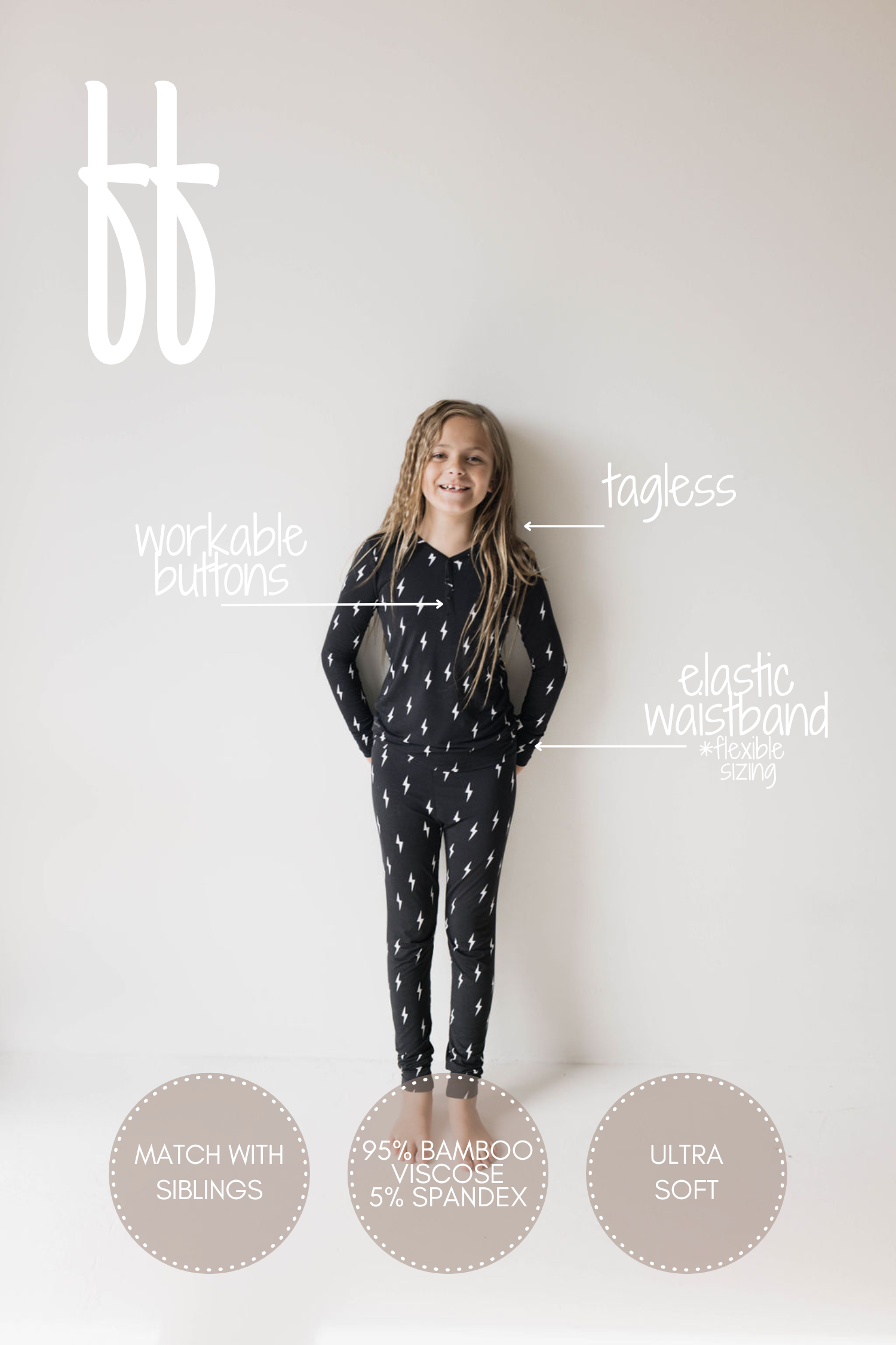 A young girl with long hair stands against a plain white background, wearing the "Bamboo Two Piece Pajamas | Midnight Lightning Bolt" by forever french baby. The ultra-soft black outfit showcases white lightning bolt patterns and features labels indicating "workable buttons," "tagless," an "elastic waistband + flexible sizing," the option to "match with siblings," and is made of 95% bamboo viscose and 5% spandex.