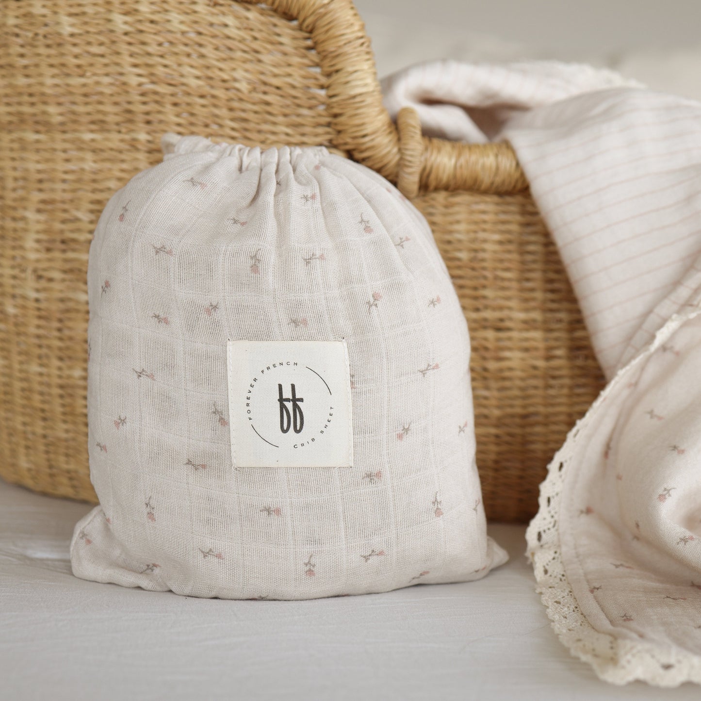 A soft, light beige Muslin Crib Sheet | Lucy by forever french baby, featuring small floral patterns and a white circular logo, is displayed in front of a woven basket with a drawstring pouch; a matching blanket lies nearby.