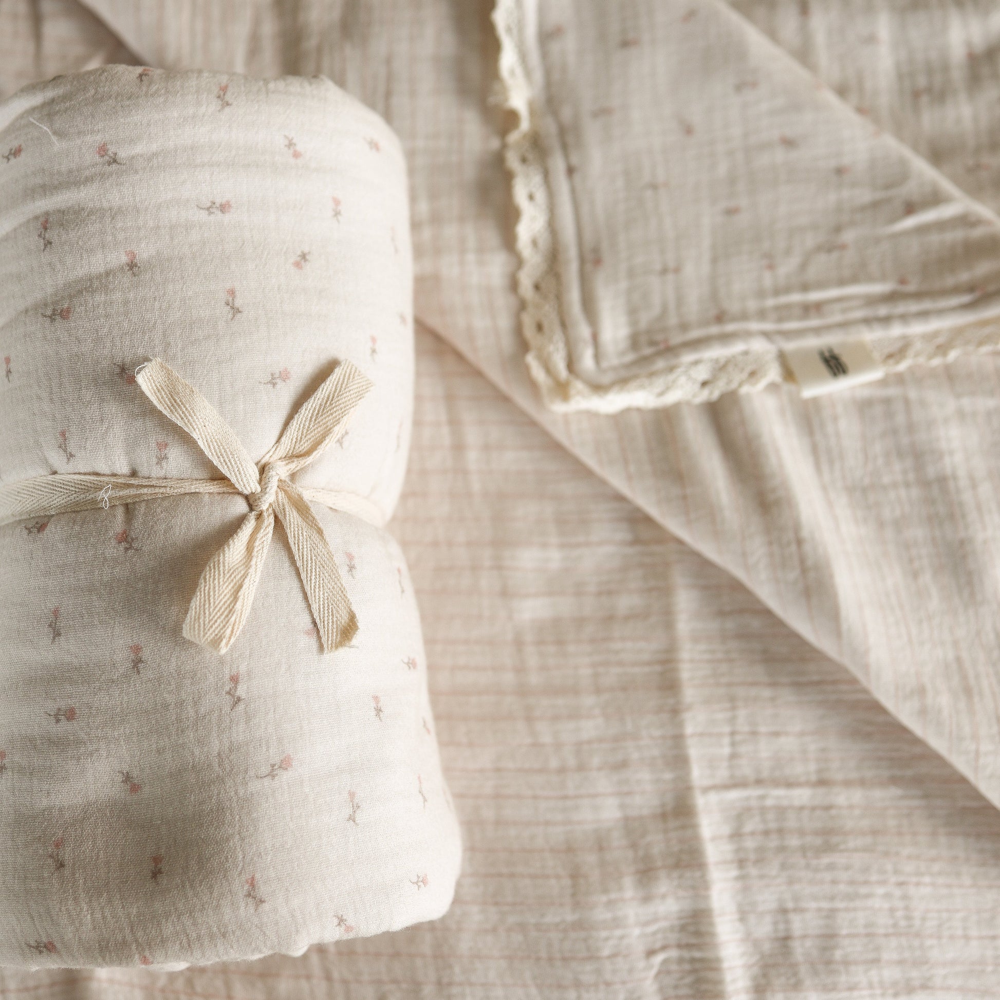 The forever french baby Muslin Quilt | Lucy features a rolled, cream-colored 100% cotton muslin blanket tied with a ribbon and an unfolded matching quilt with lace trim, both adorned with a delicate small floral pattern.