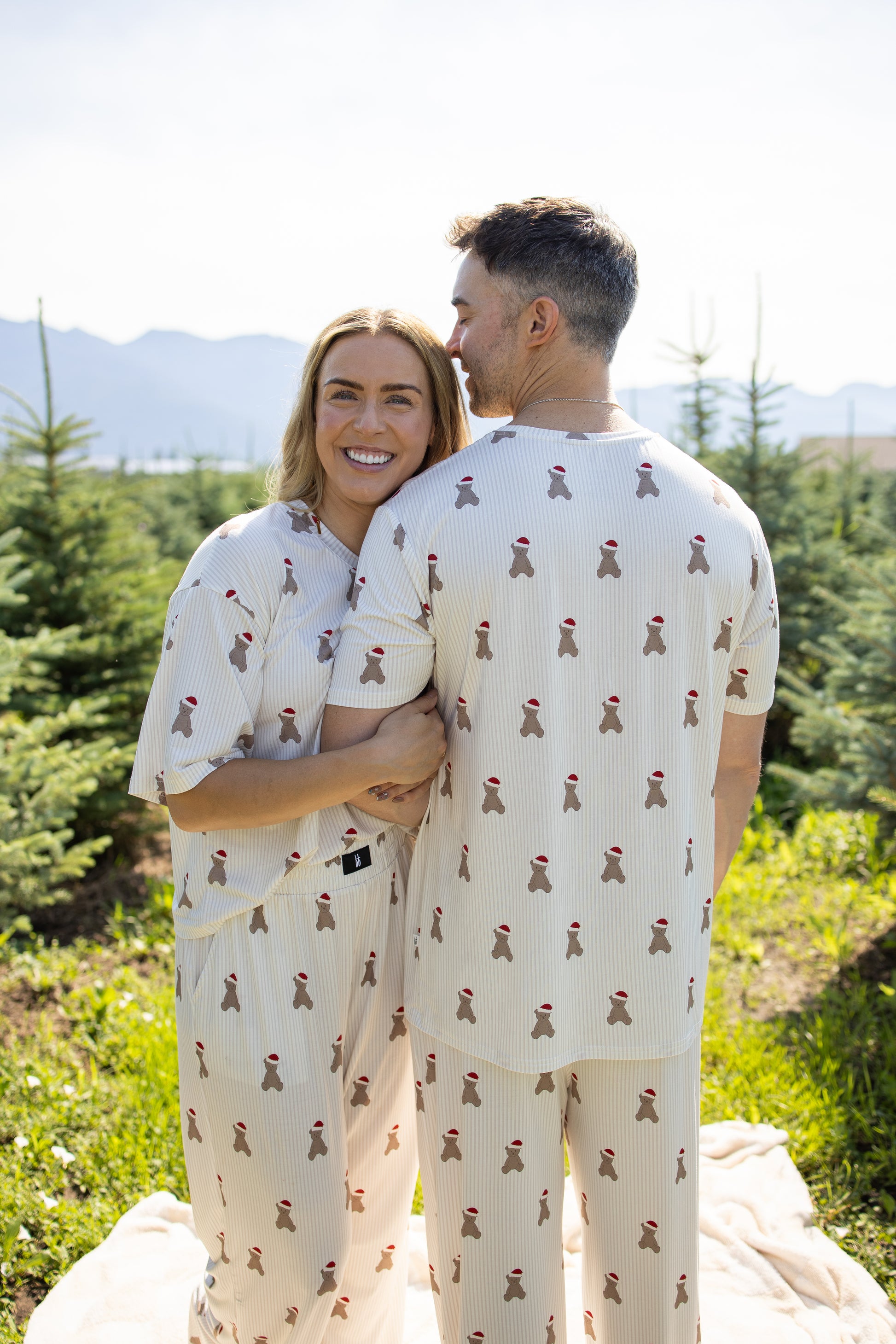 Two people stand close together outdoors, bathed in sunlight and surrounded by small pine trees, both wearing matching forever french baby Women's Boxy Tee Lounge Sets in the Santa Teddy pattern—breathable loungewear featuring cream fabric adorned with a playful red teddy bear design.