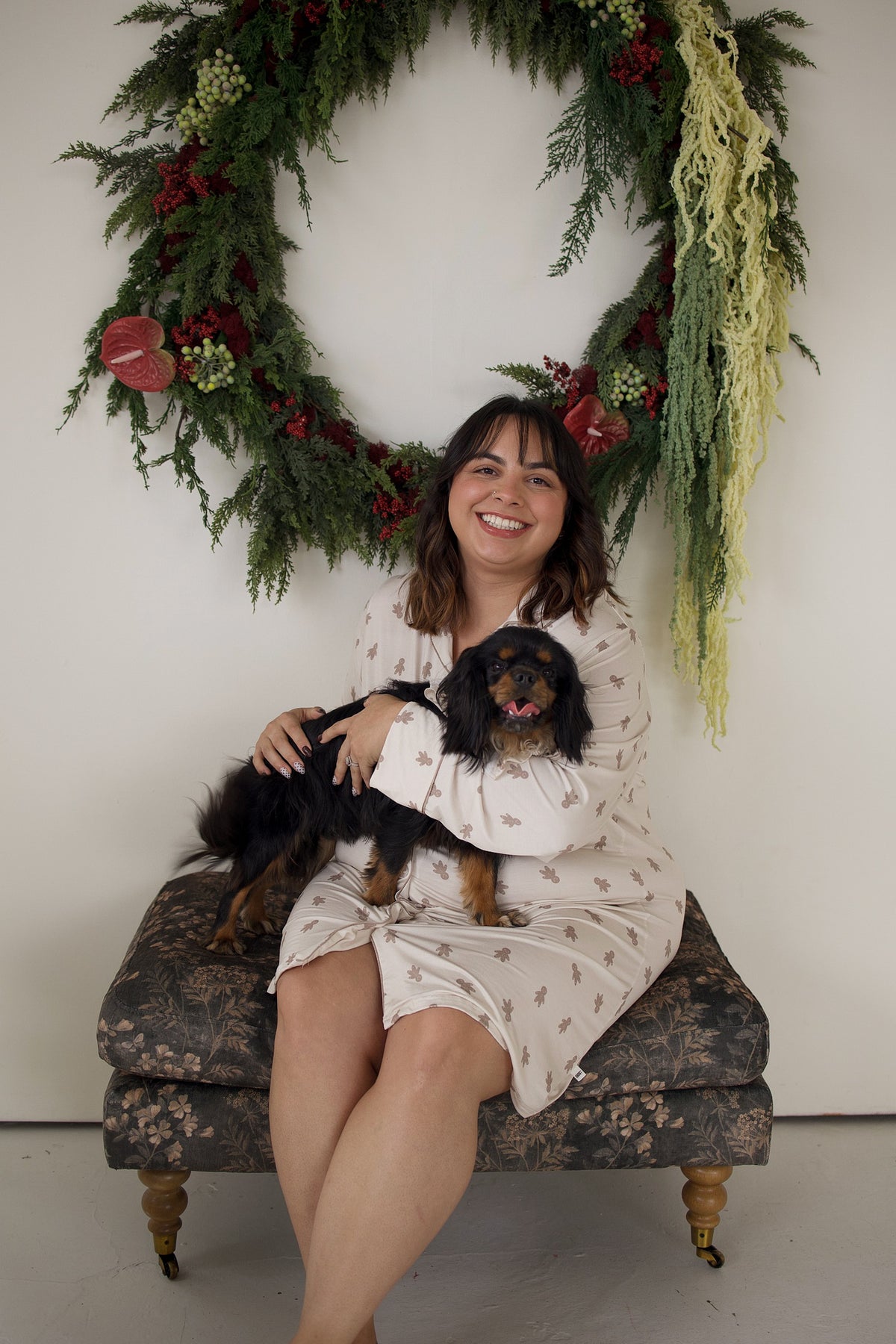 A woman wearing a Women’s Bamboo Sleeping Dress | Gingersnap sits on a patterned ottoman holding a small black and tan dog in front of a holiday wreath.