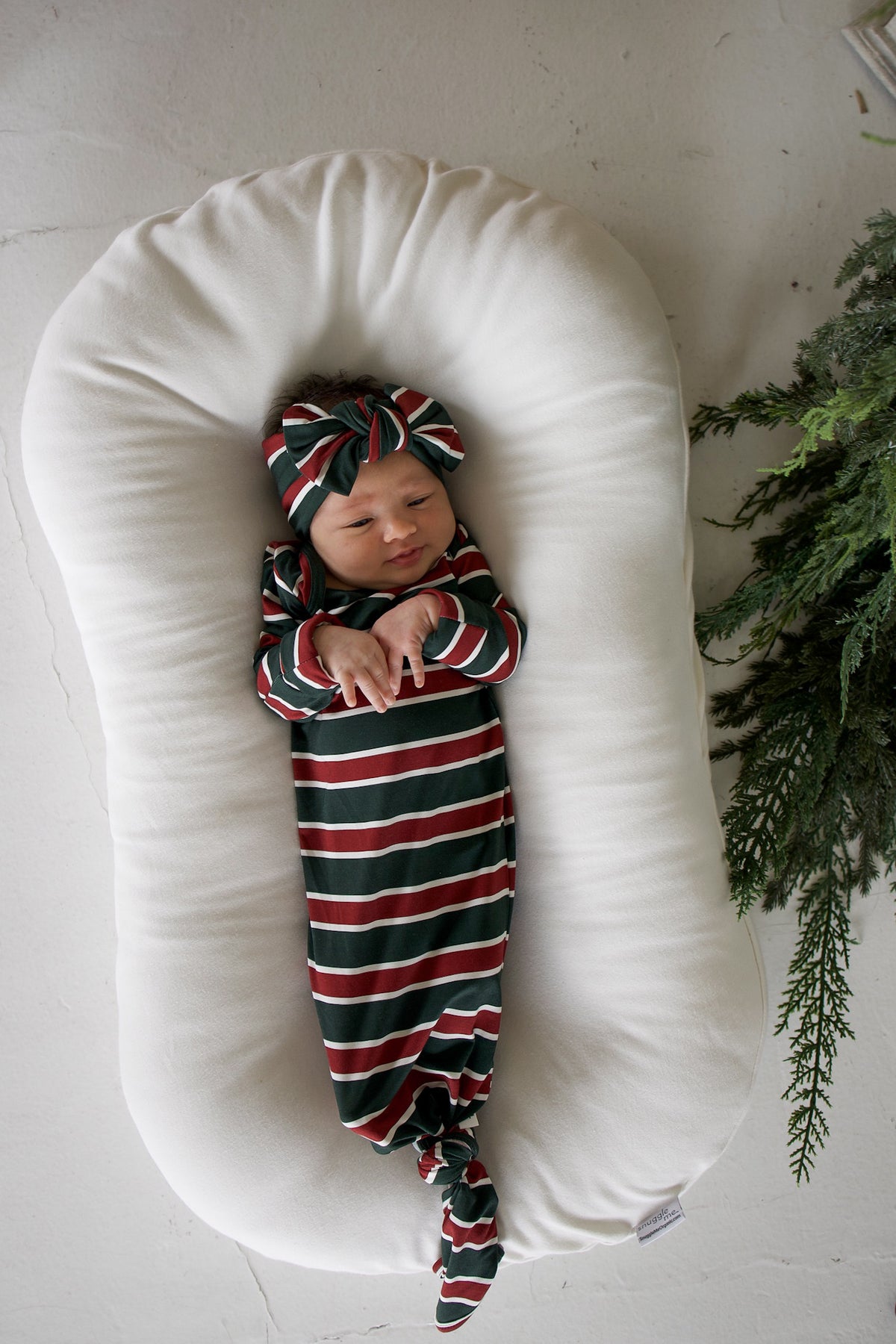 A newborn wearing a Bamboo Knotted Gown | Holiday Stripe with a matching bow headband, resting in a white infant lounger.