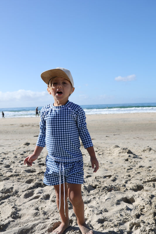 A young child models forever french baby's Child Board Short in Blue Gingham, standing barefoot on a sandy beach with the ocean and blue sky behind.