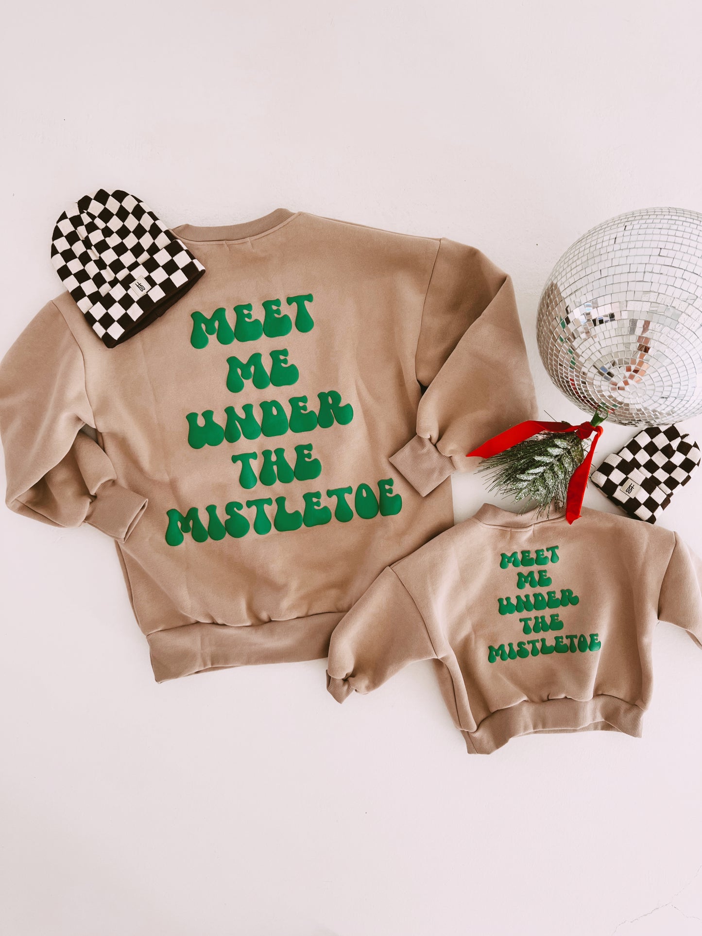 Two beige festive "Meet Me Under The Mistletoe" sweatshirts from forever french baby are displayed flat against a white background. One is adult-sized, and the other is child-sized, both featuring green text. A black-and-white checkered beanie and a sprig of greenery with a red ribbon complete this delightful holiday apparel set.