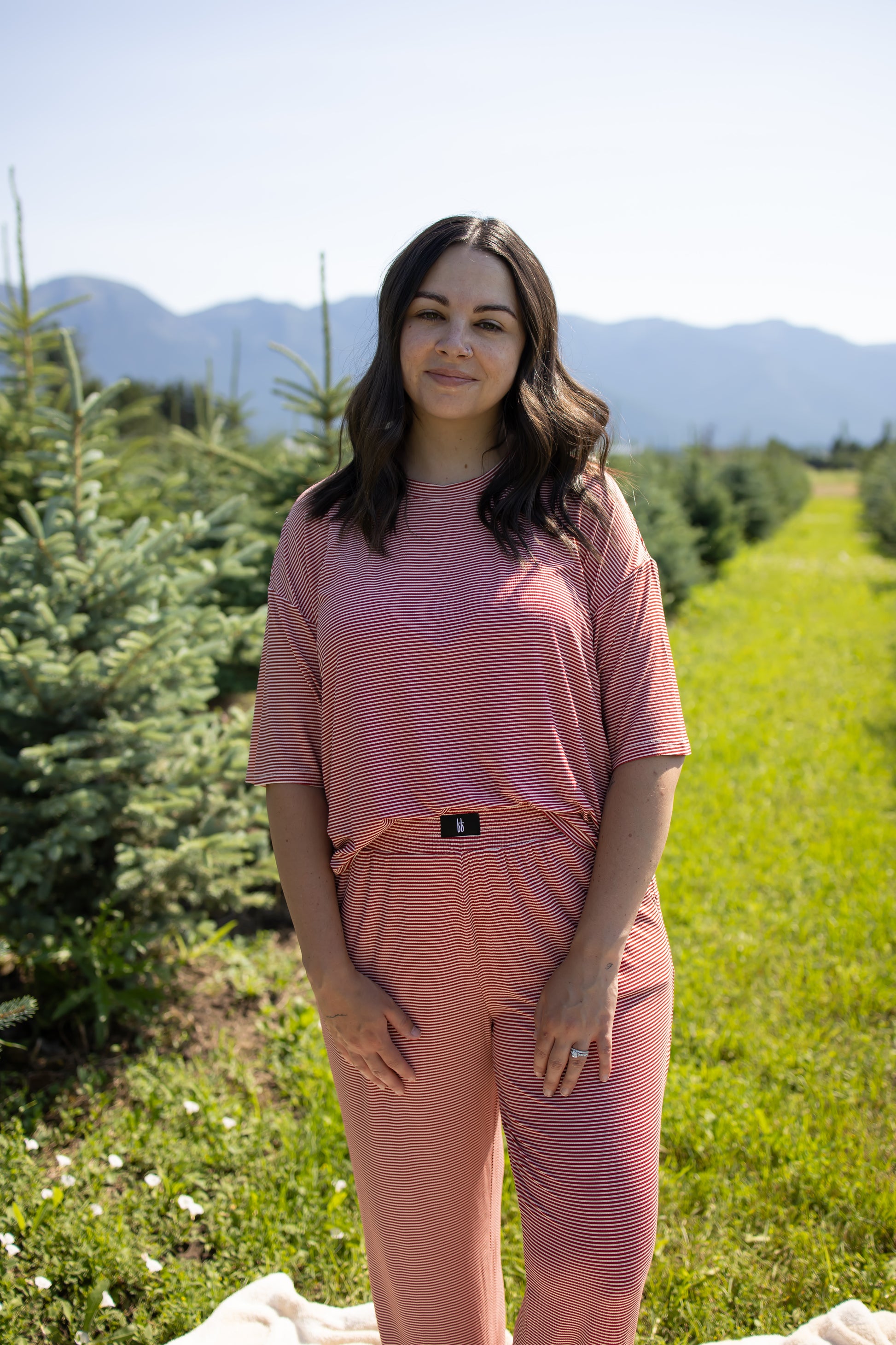 A woman with wavy brown hair stands in a field of evergreen trees, smiling softly at the camera. She is wearing the Women's Boxy Tee Lounge Set in Peppermint Stripe Ribbed from forever french baby. Mountains and a clear blue sky provide a scenic backdrop.