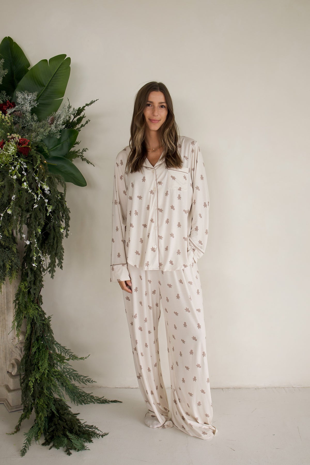 A woman wears a light-colored Women’s Bamboo Button Up Lounge Set | Gingersnap, featuring a long-sleeve button-up top and matching wide leg lounge pants with a small gingerbread man print.