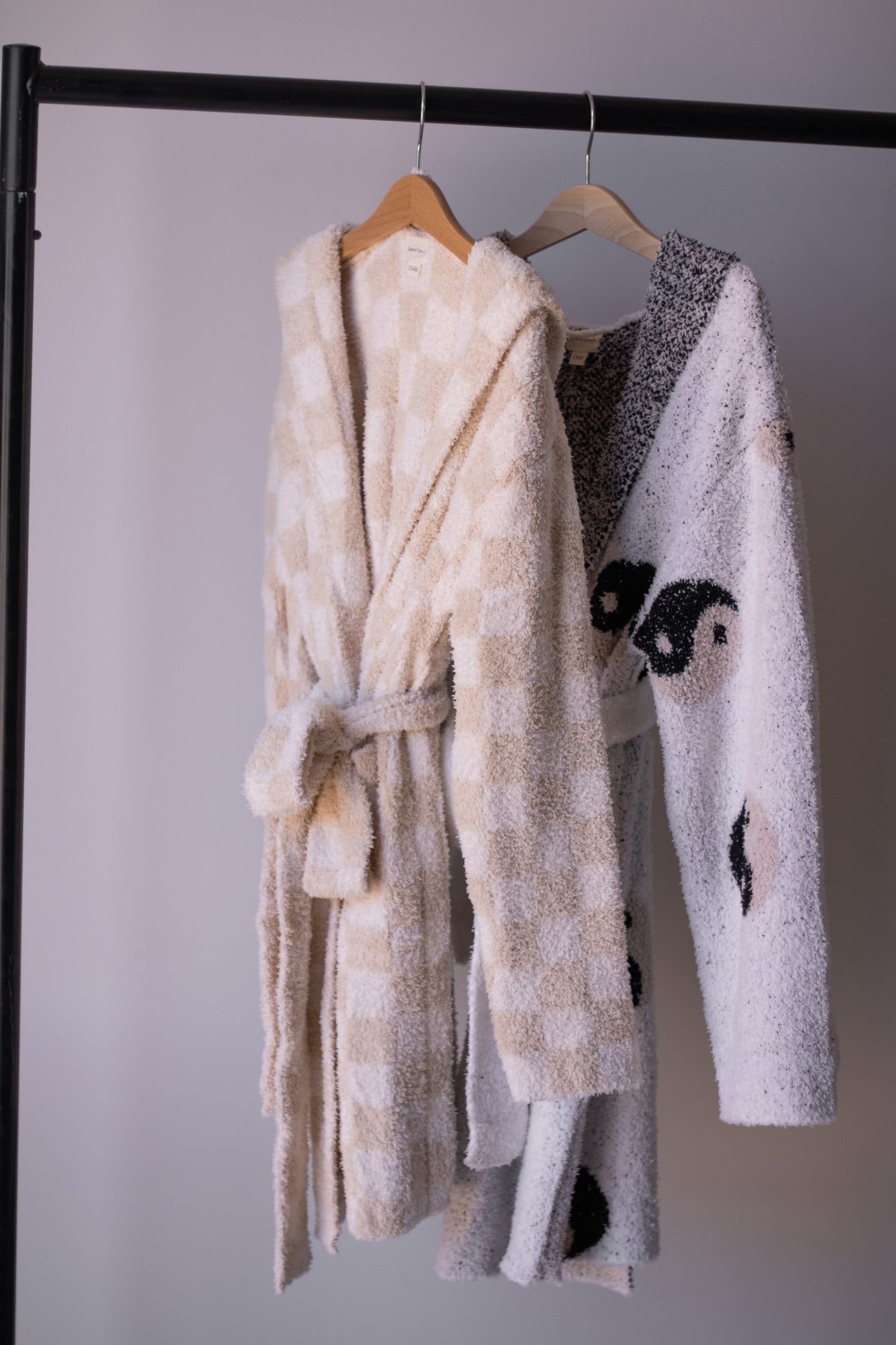 Two forever french baby bathrobes hang on a black rack: the Adult Robe | Cream Checkerboard in cream and white checks, and a white microfiber robe with a black yin-yang motif. Both are displayed on wooden hangers.
