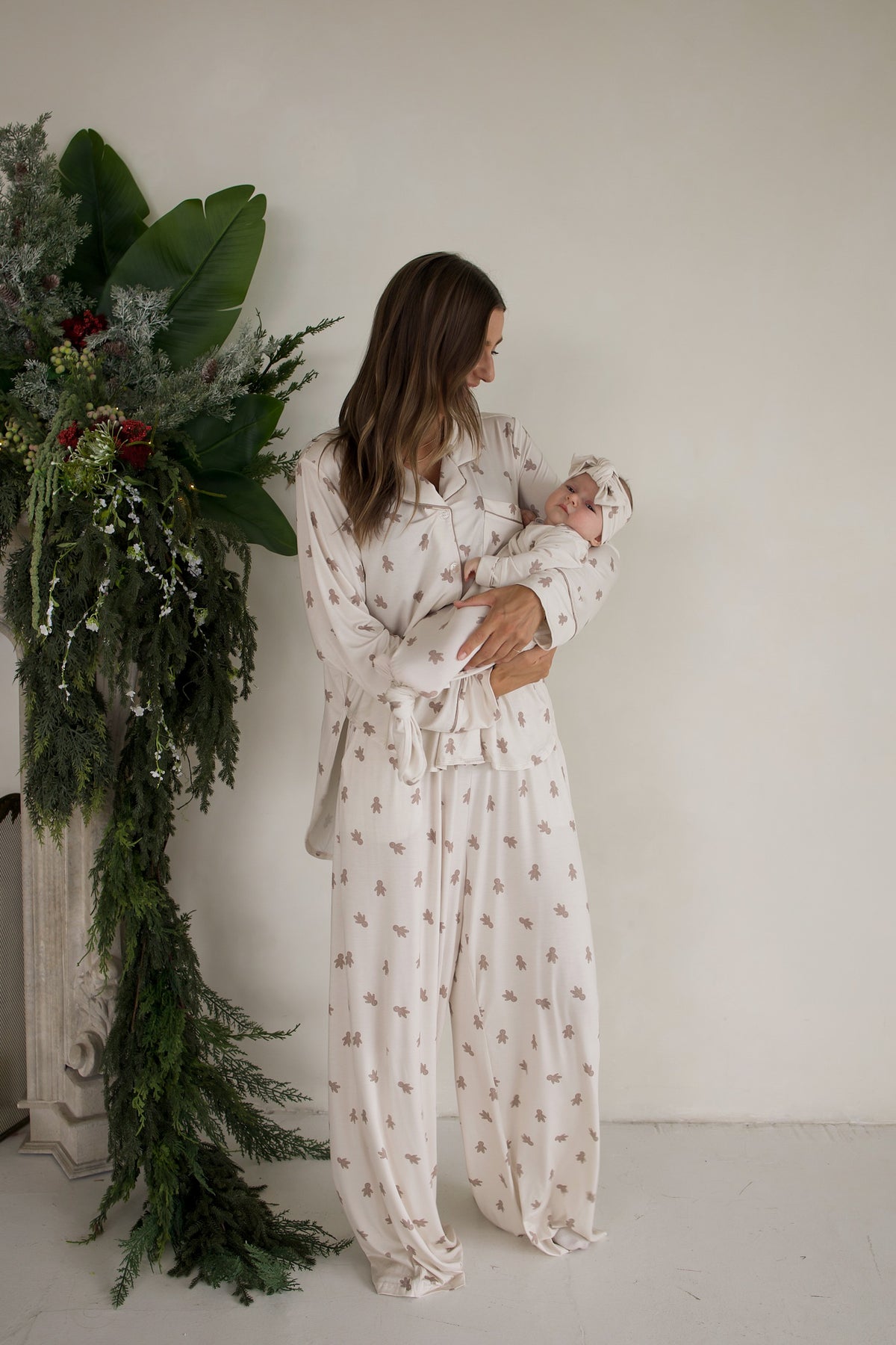 A woman in a Women’s Bamboo Button Up Lounge Set | Gingersnap holds a baby dressed in a matching gingerbread man print outfit and headband.
