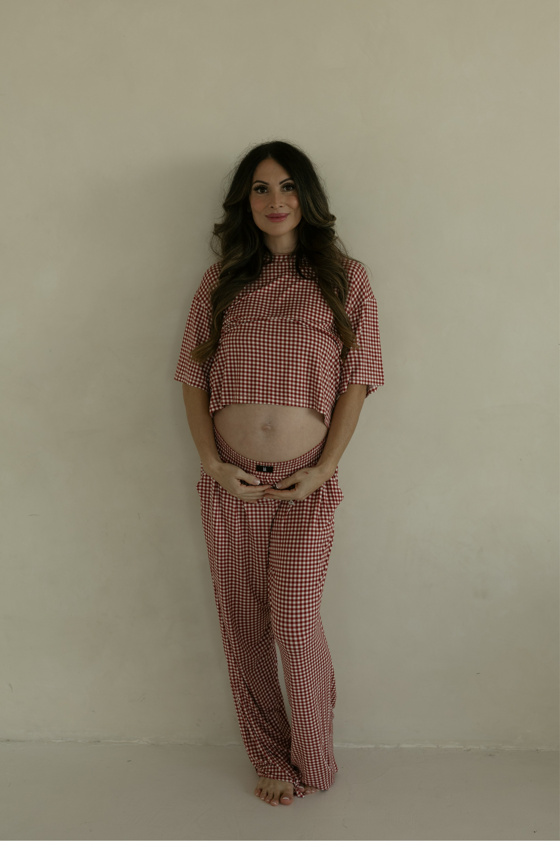 A pregnant woman stands barefoot, smiling and holding her baby bump in the forever french baby Women's Boxy Tee Lounge Set in Red Gingham, radiating comfort in these hypoallergenic pajamas.