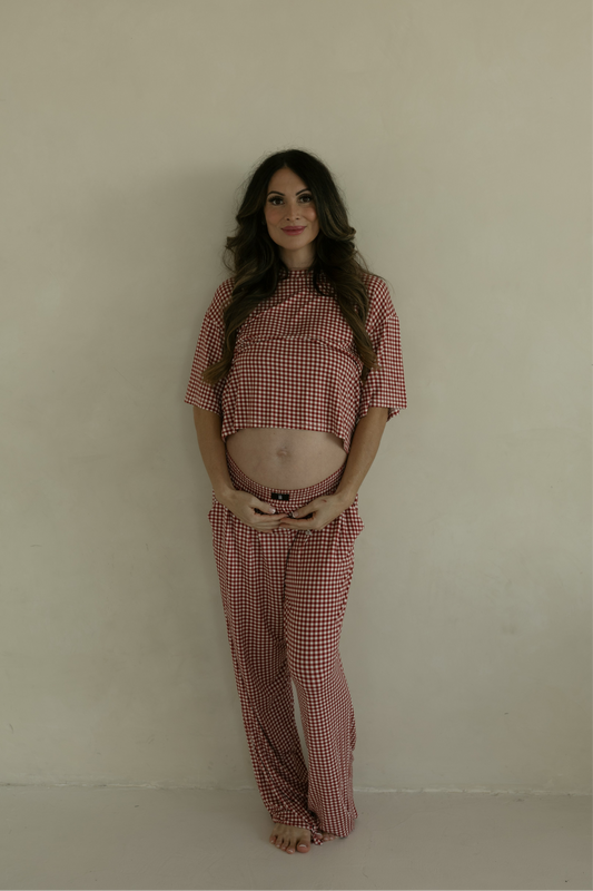 A pregnant woman stands barefoot, smiling and holding her baby bump in the forever french baby Women's Boxy Tee Lounge Set in Red Gingham, radiating comfort in these hypoallergenic pajamas.